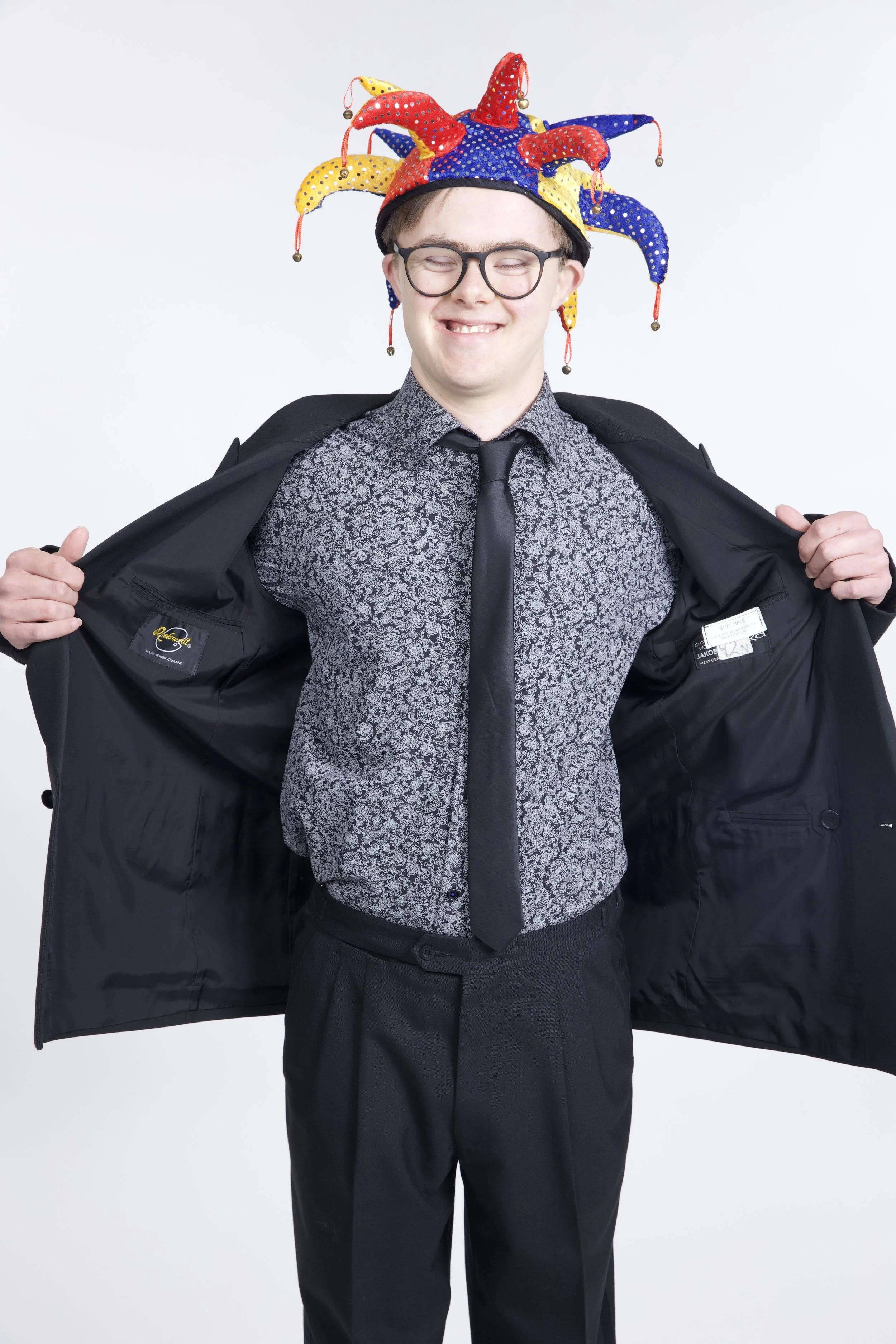 Alex is wearing a suit and tie, and a brightly coloured jester's hat with bells on it. He wears glasses, and is grinning.