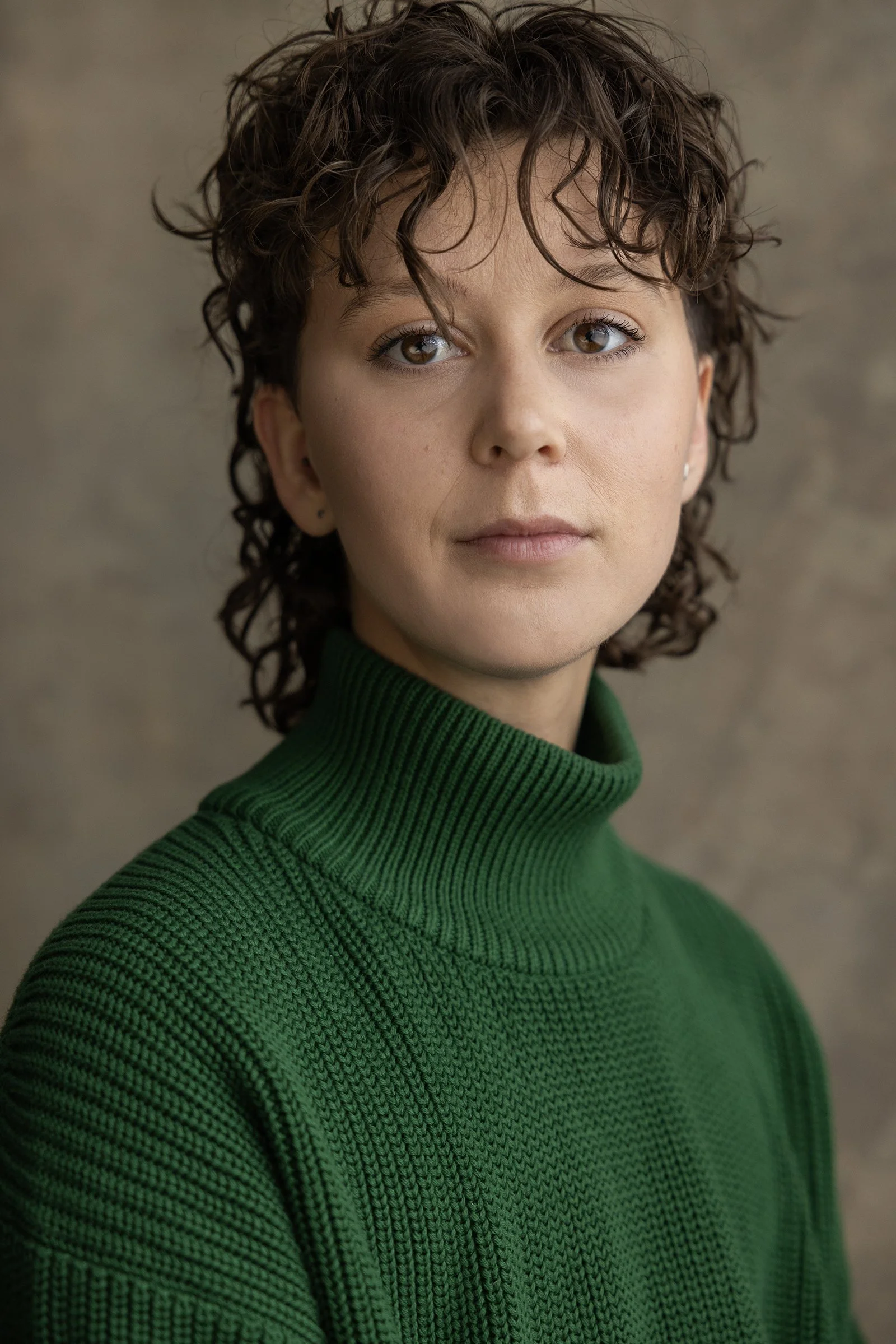 Stevie has curly brown hair in a mullet shape. They have brown eyes, and are wearing a green turtleneck sweater.