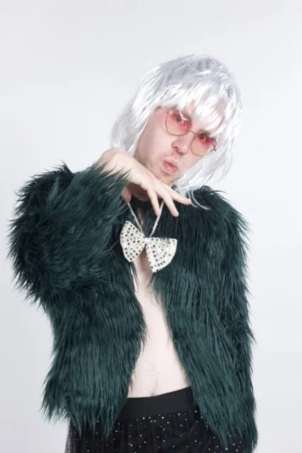 Nick is wearing a white fringed wig with pink sunglasses, a green fluffy jacket, and a gold bow tie. He stands with his hand underneath his chin, with a cheeky facial expression.