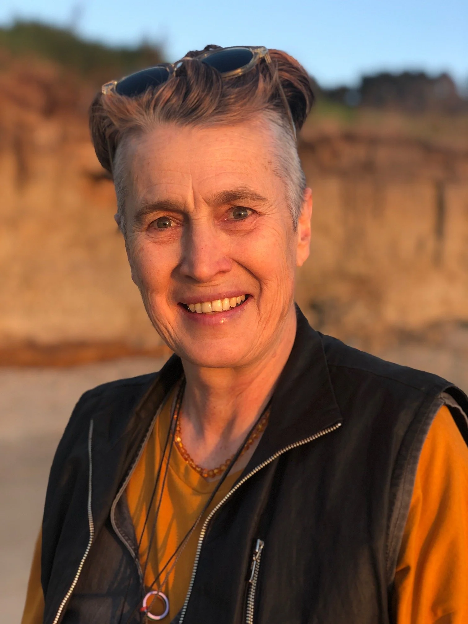 Madeline has short grey hair with shaved sides, and is wearing sunglasses on her head and smiling.