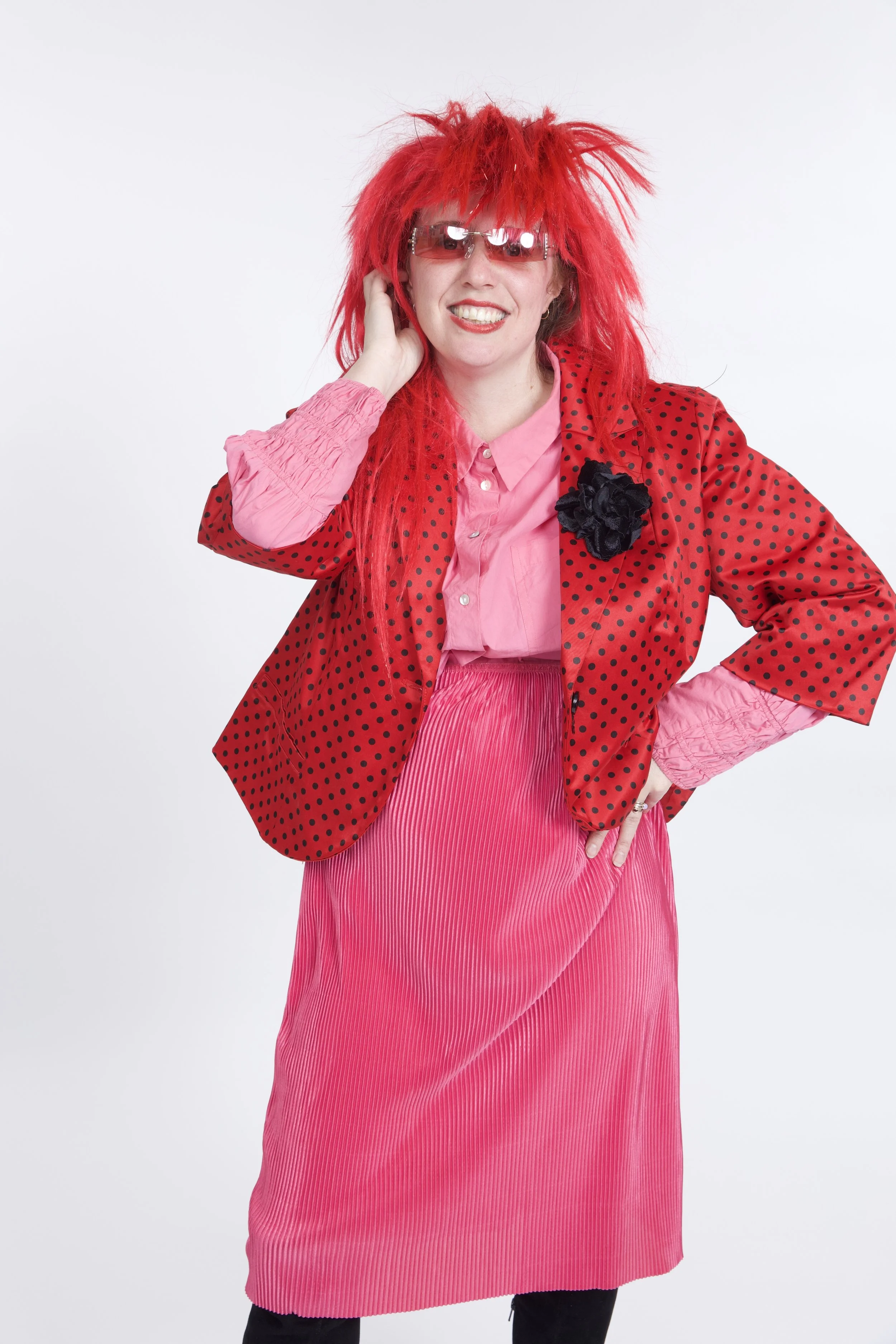 Tess is wearing an outfit out of entirely pink and red. She is wearing a red wig and red-tinted sunglasses, with red lipstick. She is grinning.