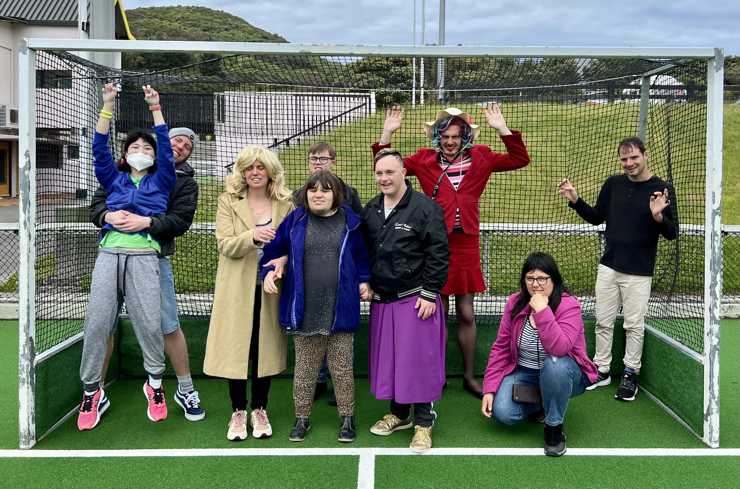 A group of 9 actors stand posed inside a hockey goal. They are wearing colourful outfits, and have joyous expressions.