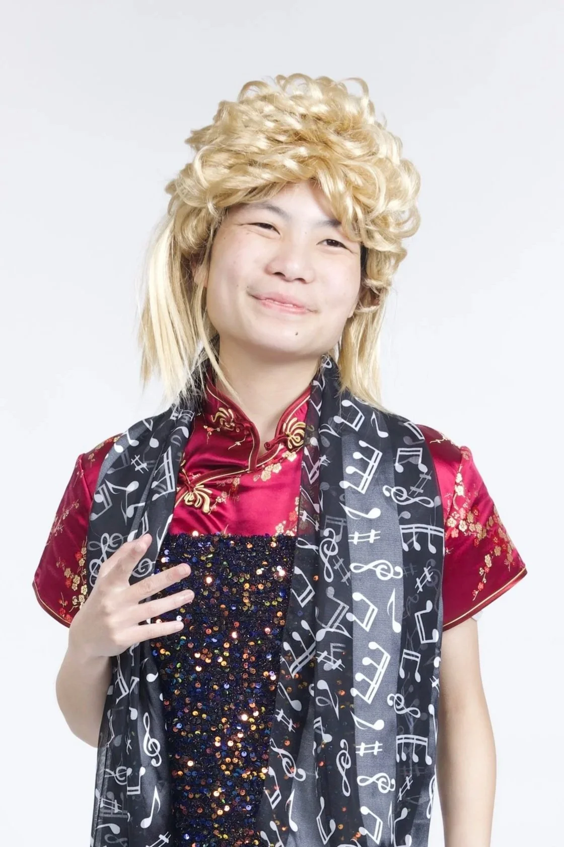 Caitlyn is wearing a blonde mullet wig, a sparkly black and red dress, and a black scarf with musical notes on it. She is smiling.
