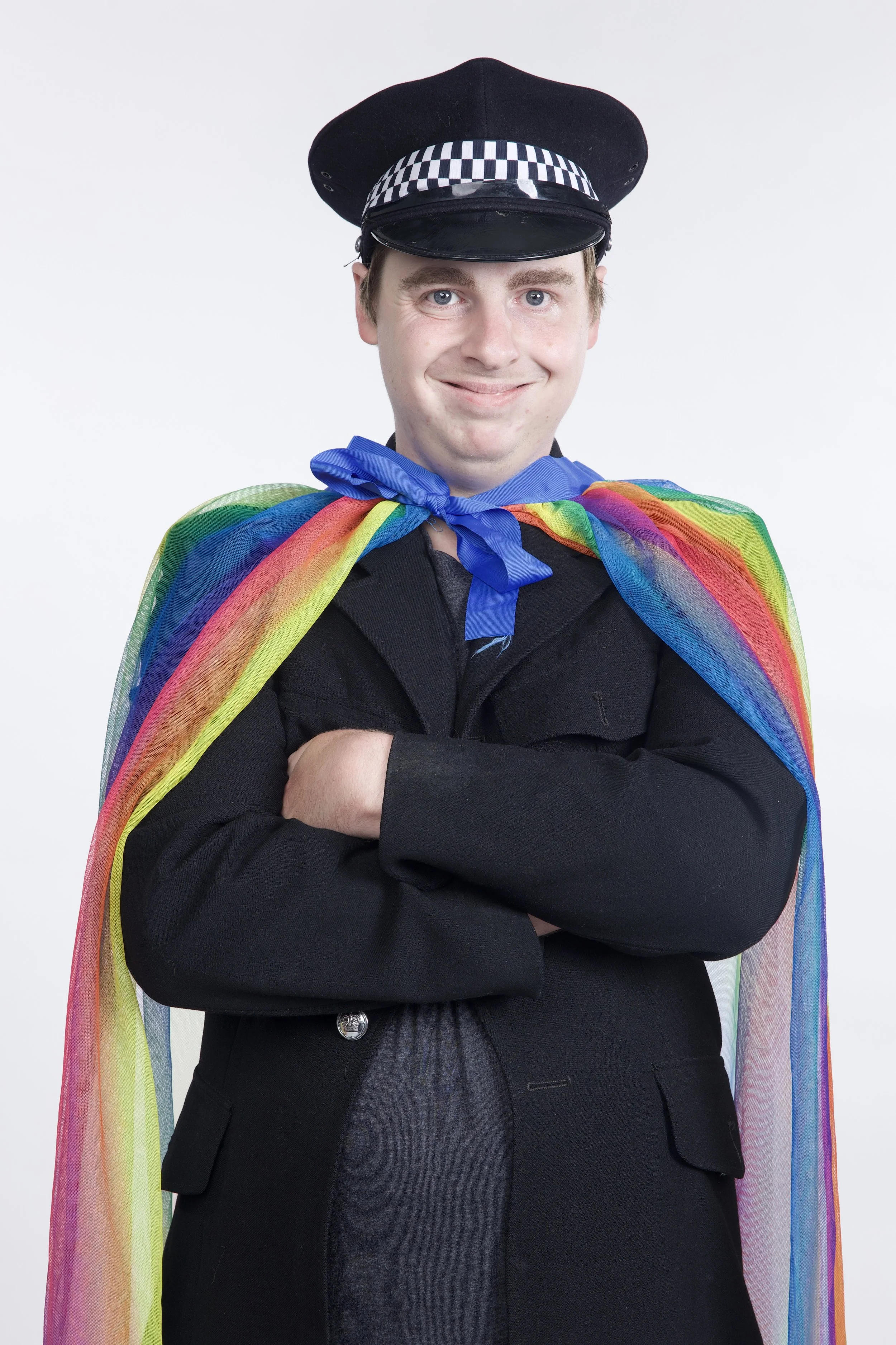 Ryan is wearing a police hat and a flowing rainbow cape over a black blazer. He is smiling and has his arms folded.