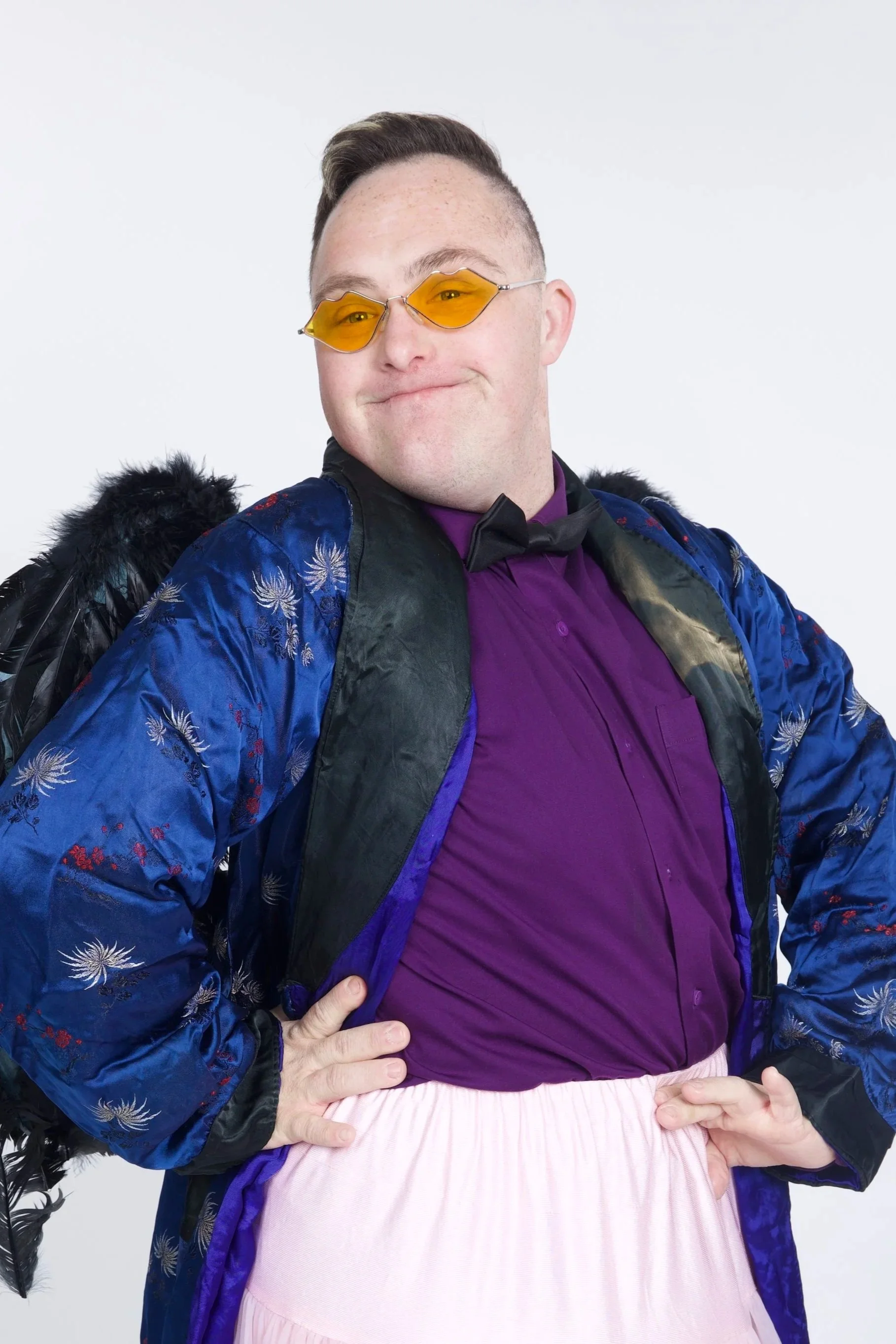 Callum has sort brown hair, and is wearing a blue jacket with a purple top and a light pink skirt. He has yellow sunglasses on, and is smiling.