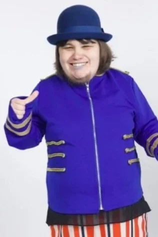 Maggie Rose is wearing a blue bowler hat and a blue jacket. She has short brown hair and a big grin.