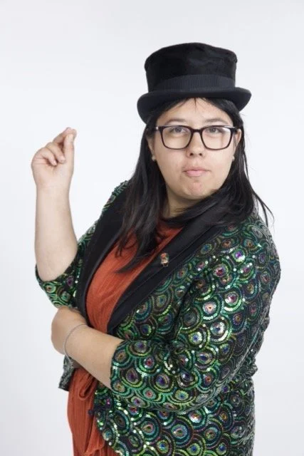 Aurora has one arm folded across her body and the other up near her face. She is wearing a top hat, and a green sparkly jacket. She wears glasses, and a focused expression.