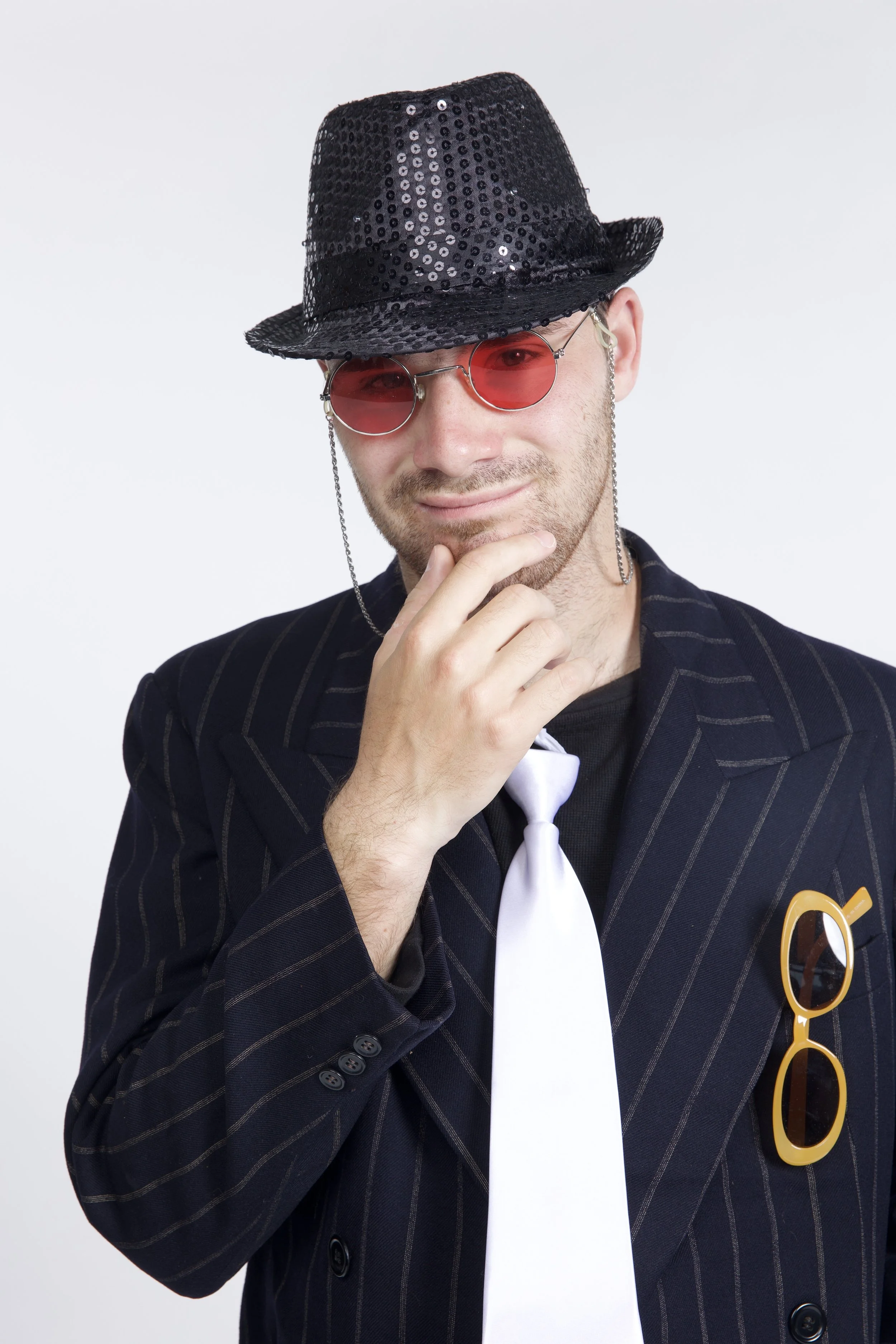 Jeremy is wearing a pinstriped suit and tie, a sparkly fedora, and red-tinted sunglasses on a chain. He has his hand to his chin in a pensive position.
