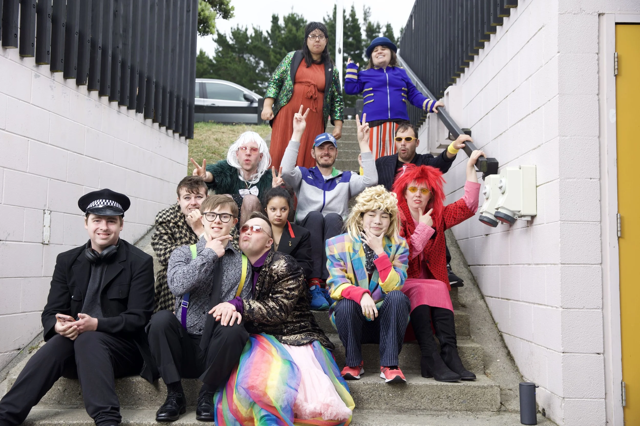 A group of 12 actors sit together on some concrete steps. They are all wearing bright colourful costumes.