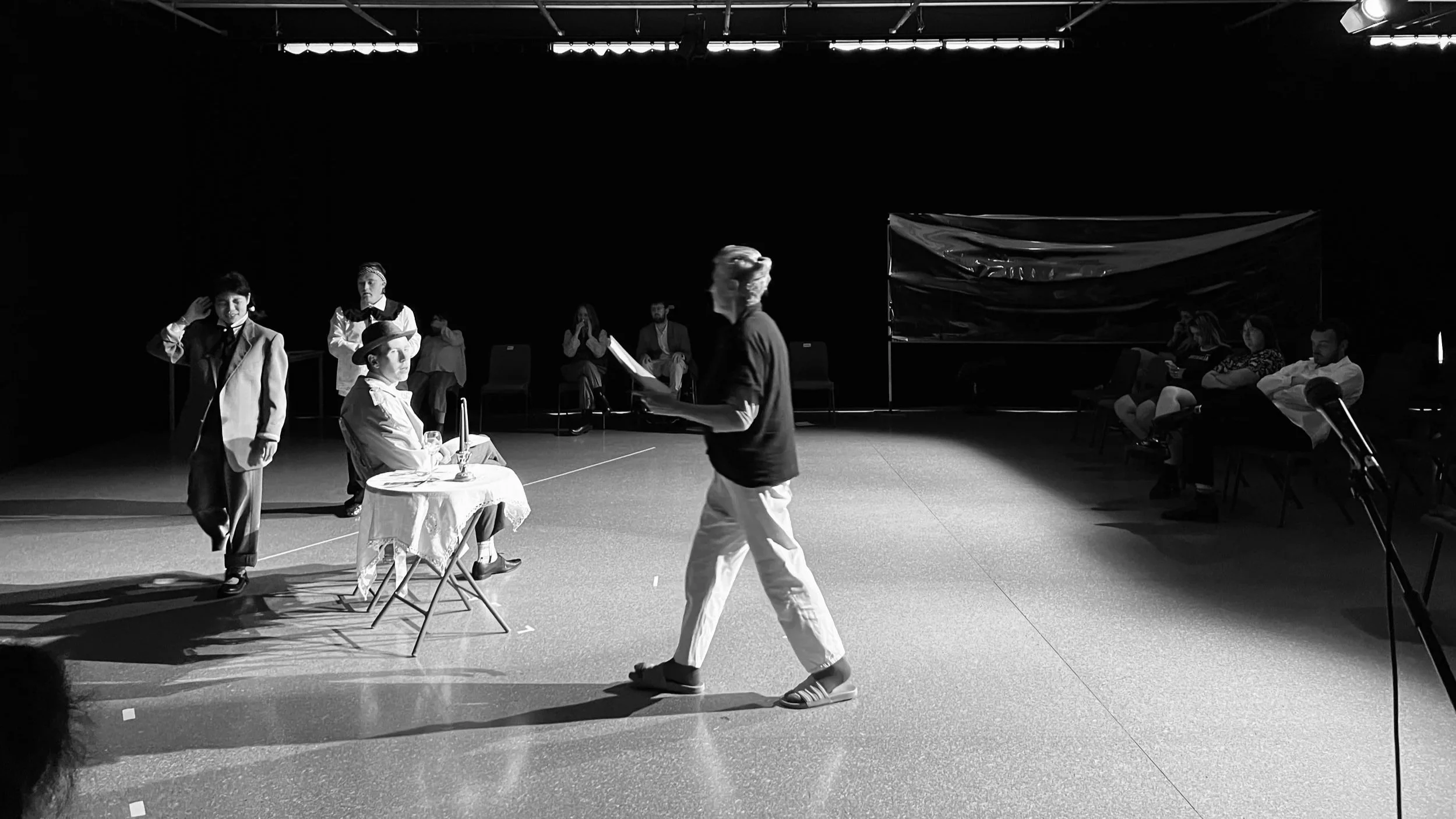A black and white photograph of a director working with three actors in a theatre space. One actor is seated at a table, and two others are walking behind. They are all wearing suits.