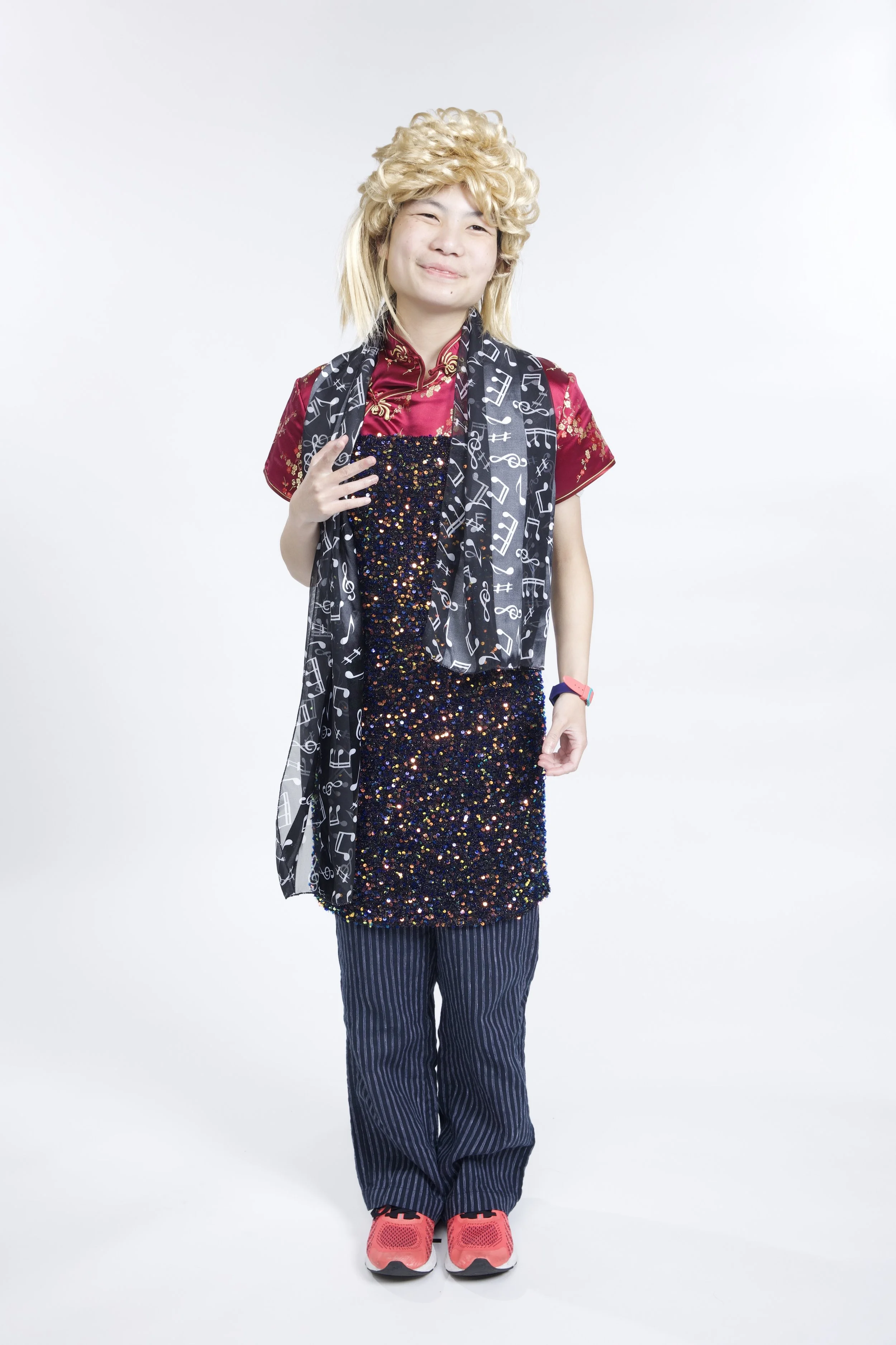 Caitlyn stands with a serene expression on her face. She is wearing a blonde curly mullet wig, a sparkly dress, and a scarf with musical notes on it.