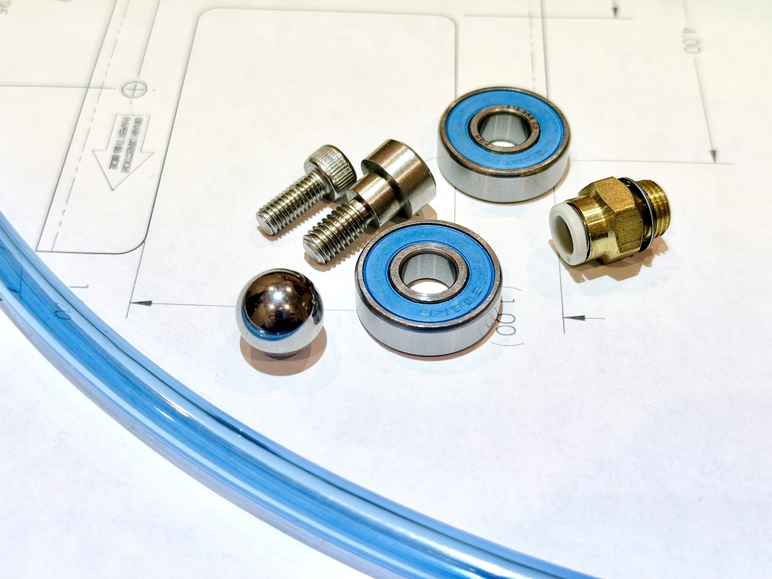 Close-up of mechanical parts including bearings, shoulder bolts, a ball bearing, a pneumatic fitting, and a tube on technical drawings.
