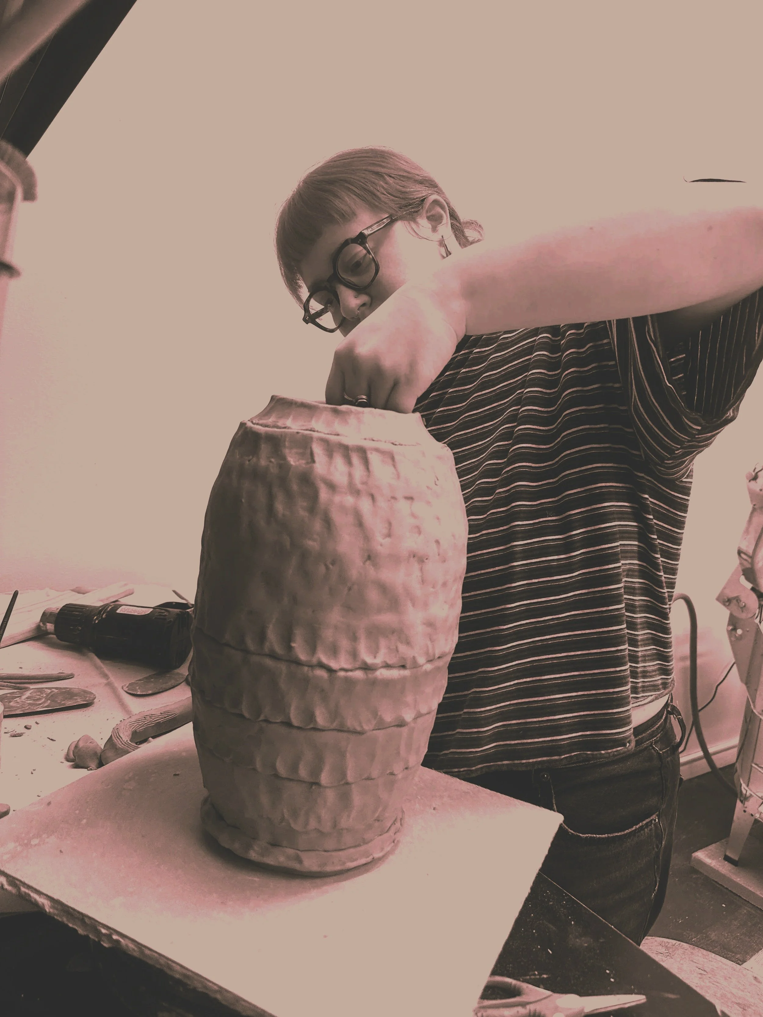 A person with glasses, striped shirt, and a ponytail working on a large ceramic vase or sculpture in a workshop. The workspace has tools and equipment on the table and around.