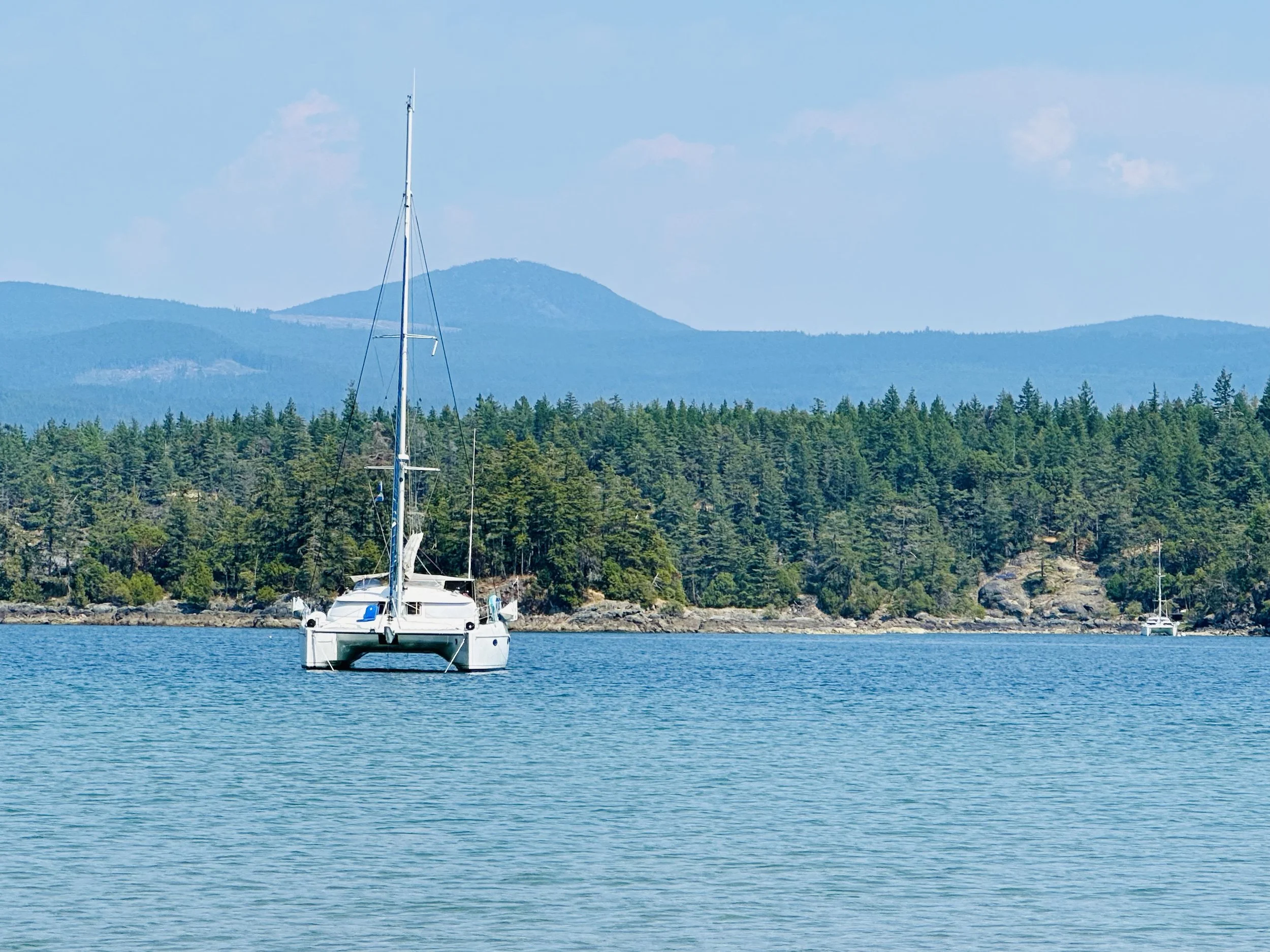 A sailboat anchored in calm blue water with a forested shoreline and mountains in the background.