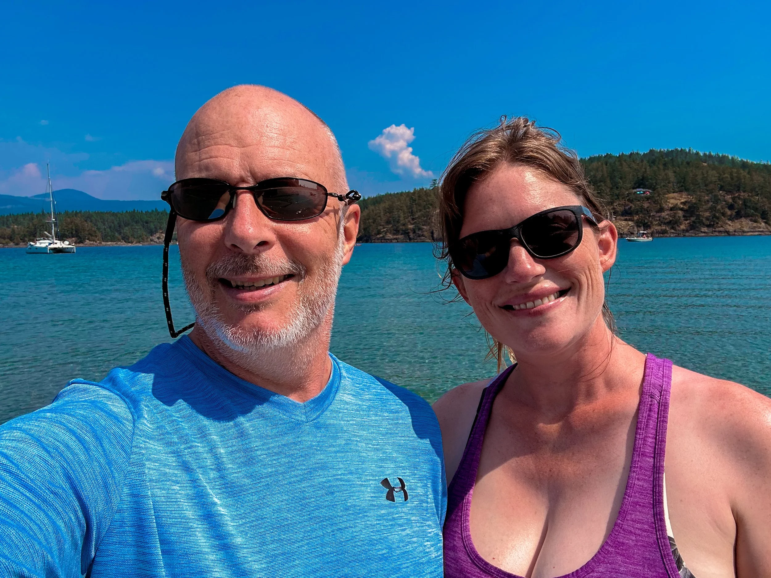 A smiling man and woman wearing sunglasses taking a selfie near a lake with sailboats and a forested shoreline under a clear blue sky.