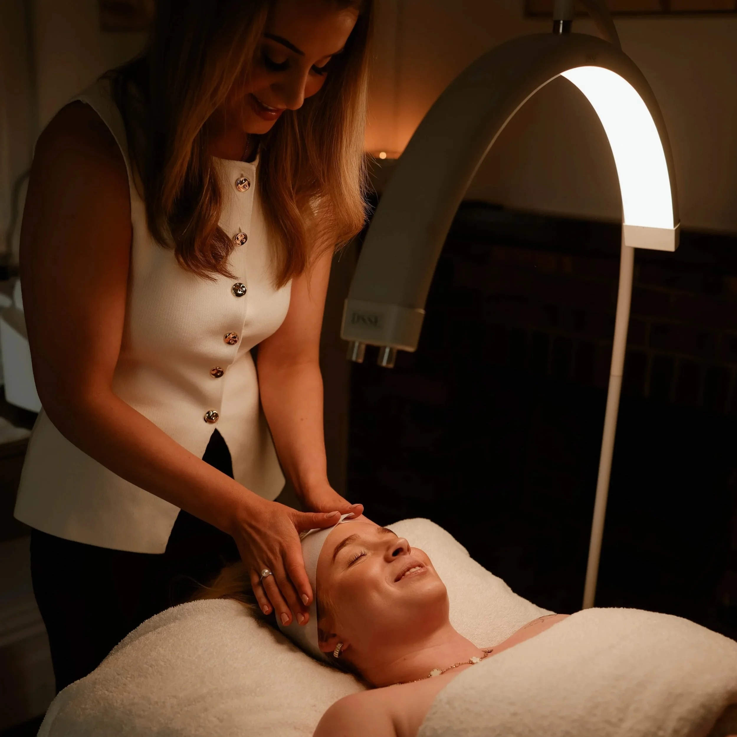 A woman receiving a facial treatment or massage in a spa-like setting with warm lighting, lying on a treatment bed, while another woman gently places her hands on her face.