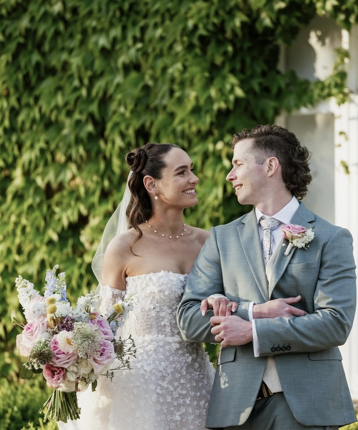 A bride and groom smiling at each other on their wedding day outdoors, with green foliage in the background. The bride is holding a bouquet of flowers and wearing a white lace wedding dress and veil. The groom is dressed in a light gray suit with a b