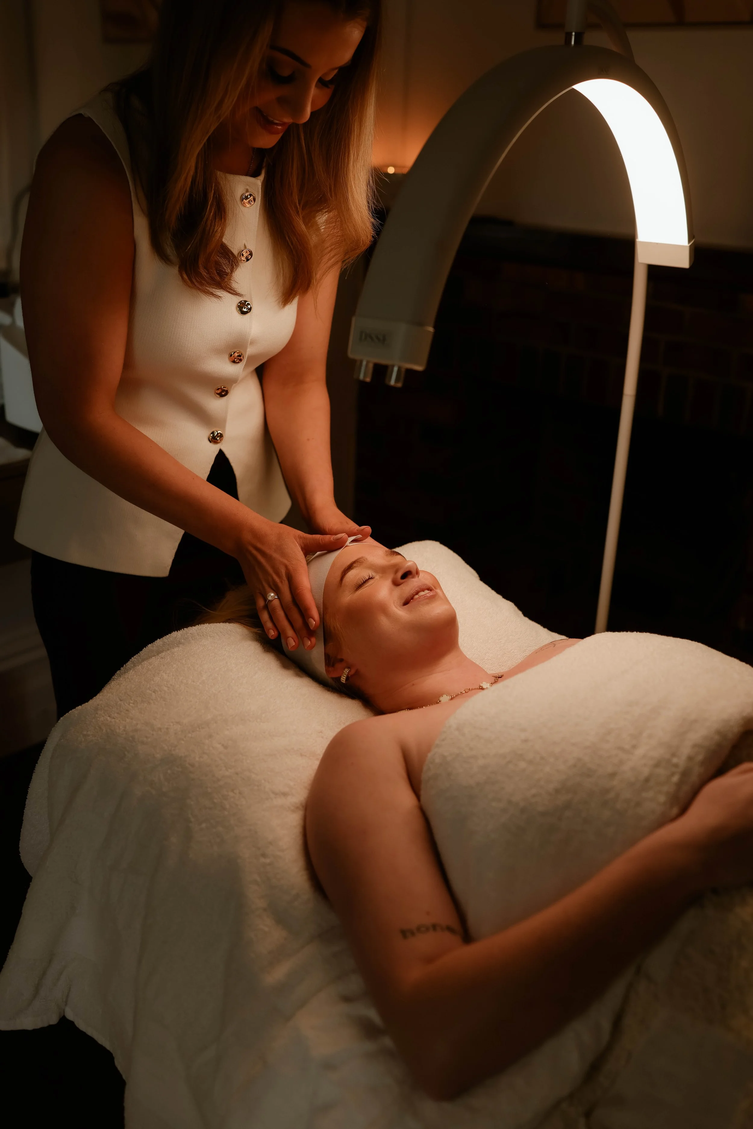 A woman lying on a treatment bed with her eyes closed and a content smile, receiving a facial treatment from a professional healthcare provider under a bright, curved medical lamp in a spa or clinic setting.