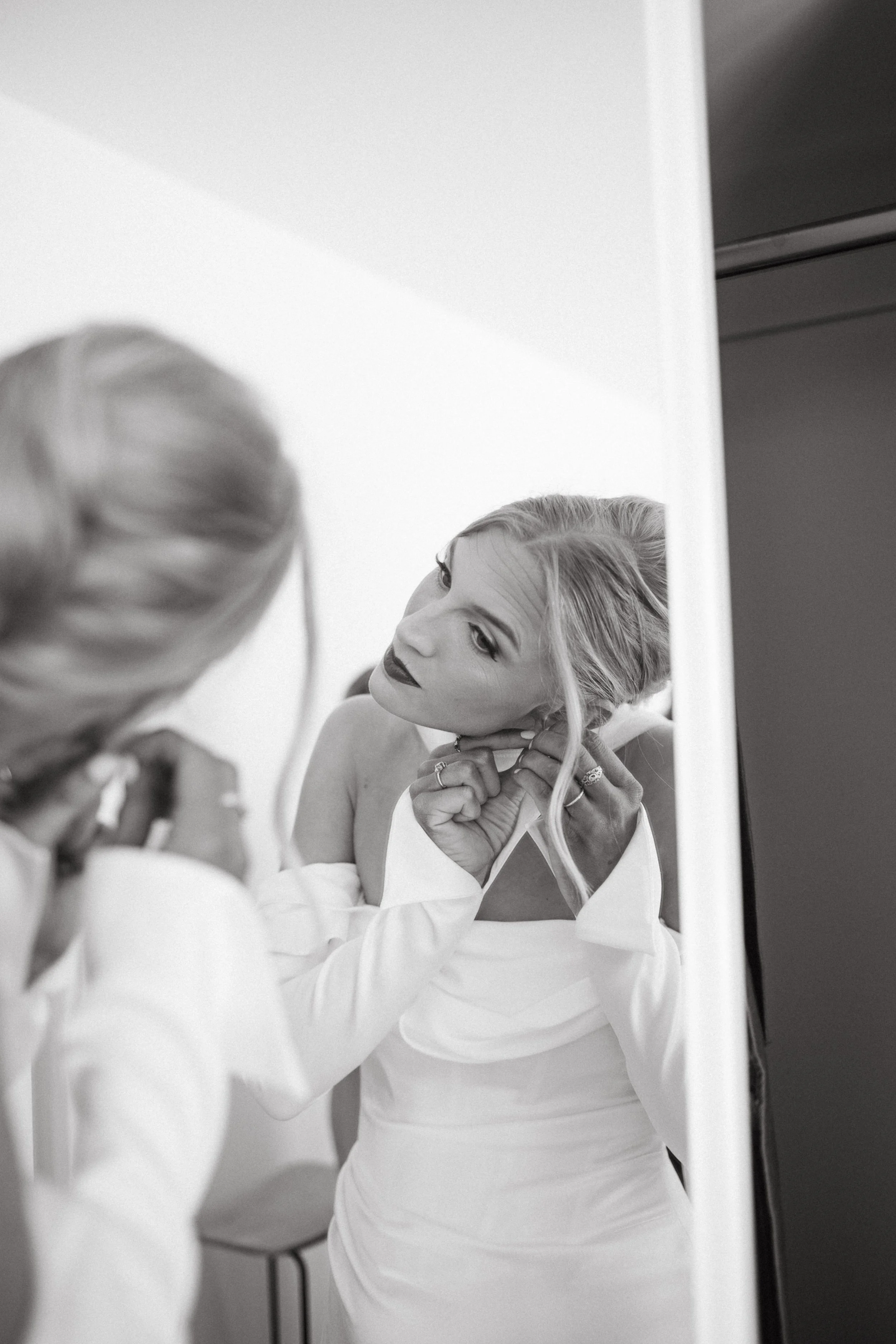 A woman with blonde hair and dark lipstick is looking at herself in a mirror while adjusting her earrings. She is wearing a white dress, and her reflection is visible.