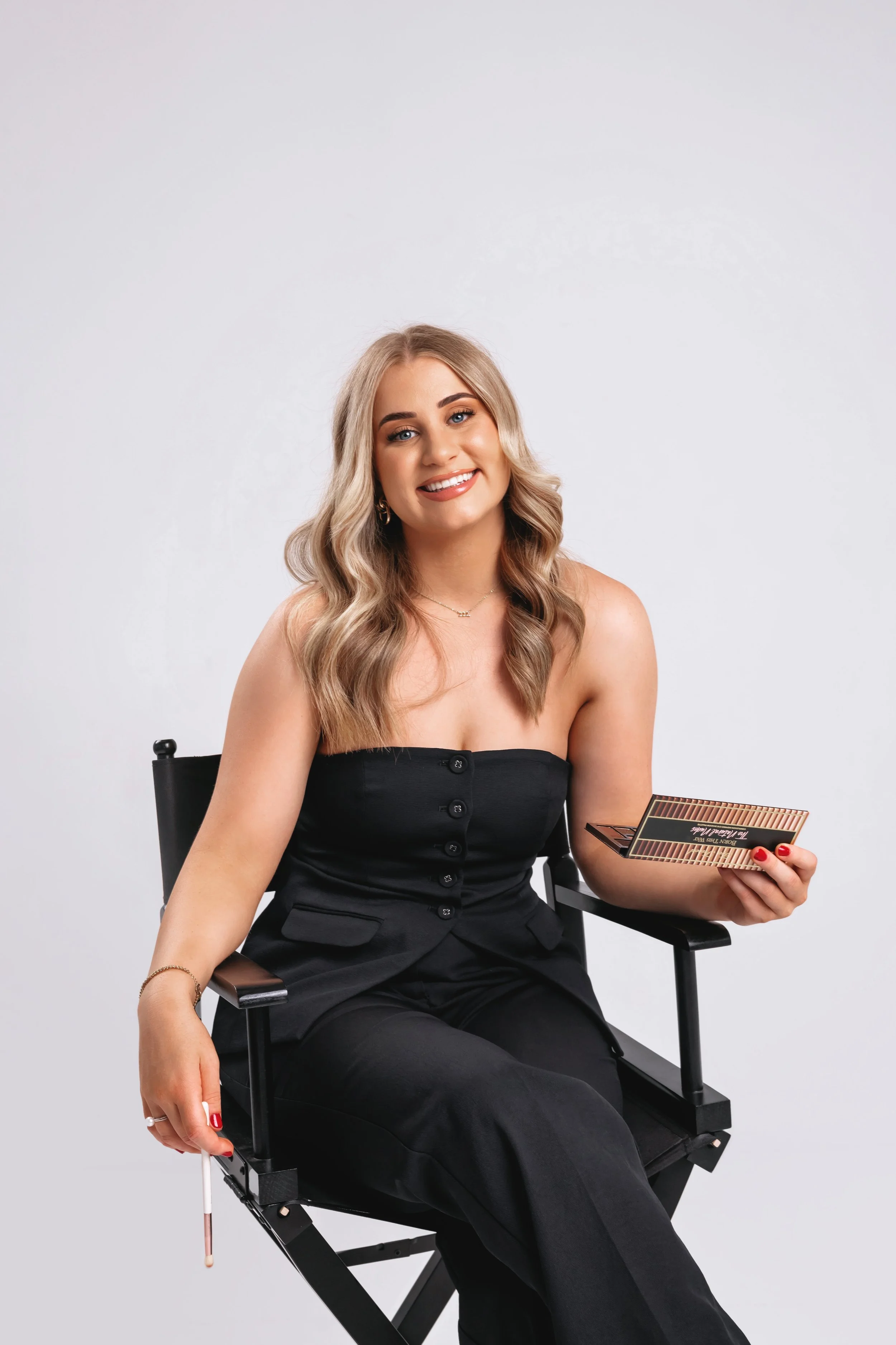 A smiling woman with blonde hair, wearing a black strapless top and black pants, sitting on a director's chair, holding an eyeshadow palette.