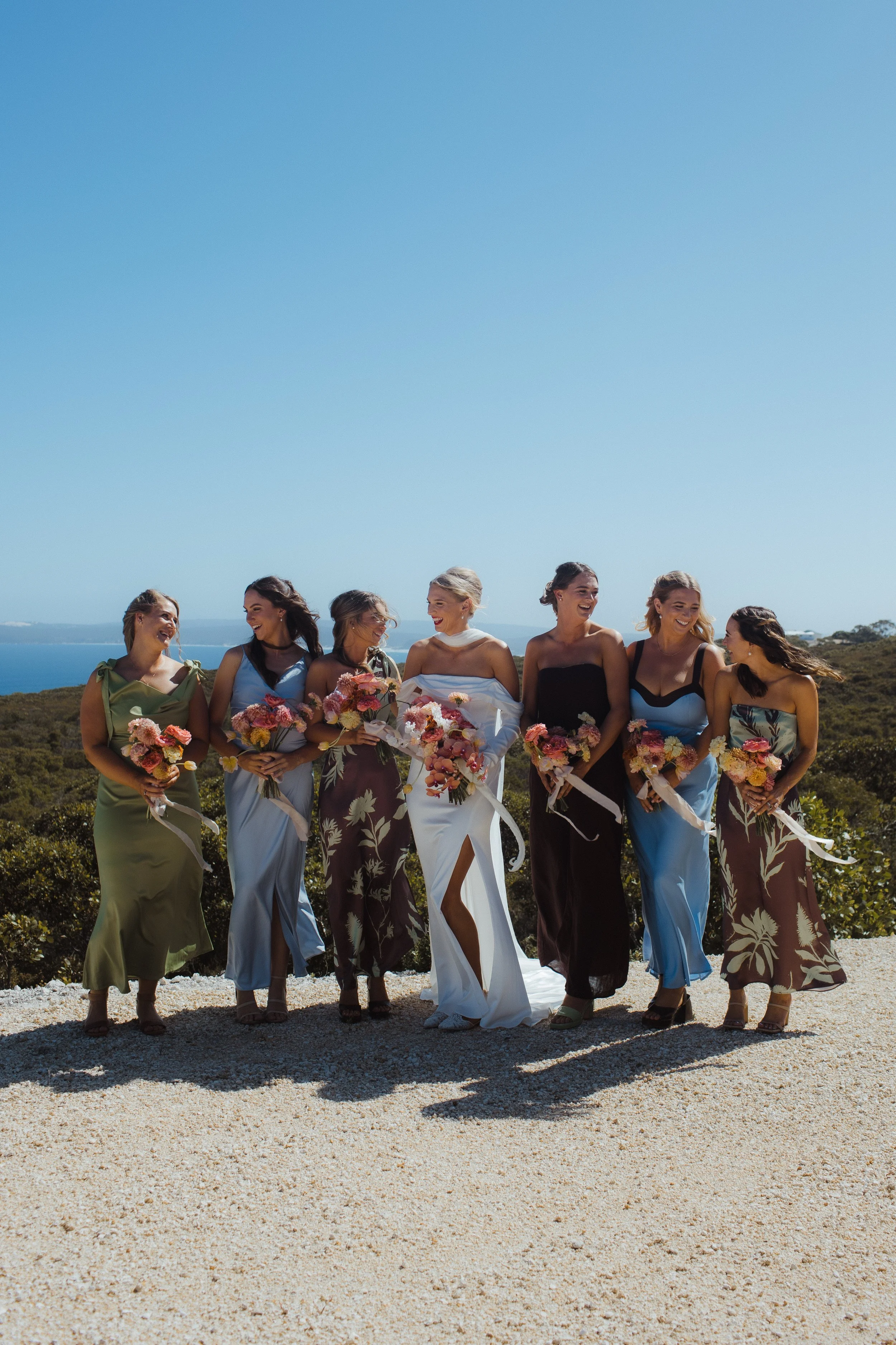 A bride and six bridesmaids in colorful dresses holding bouquets, standing outdoors on a sunny day with greenery and ocean in the background.