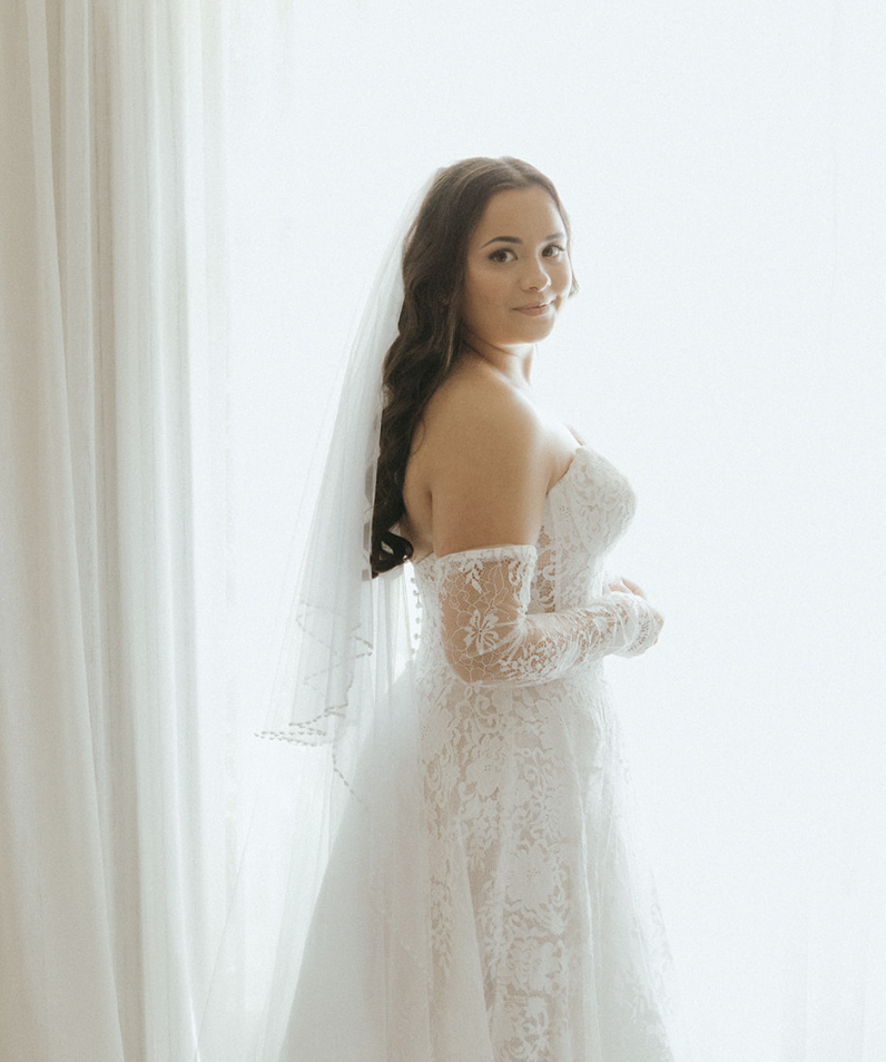 A bride in a lace wedding dress with long sleeves, standing near a window with curtains, looking at the camera with a slight smile.