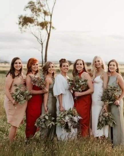 Group of seven women standing outdoors on grass, holding bouquets, dressed in formal dresses, smiling for the camera, with a tree and cloudy sky in the background.