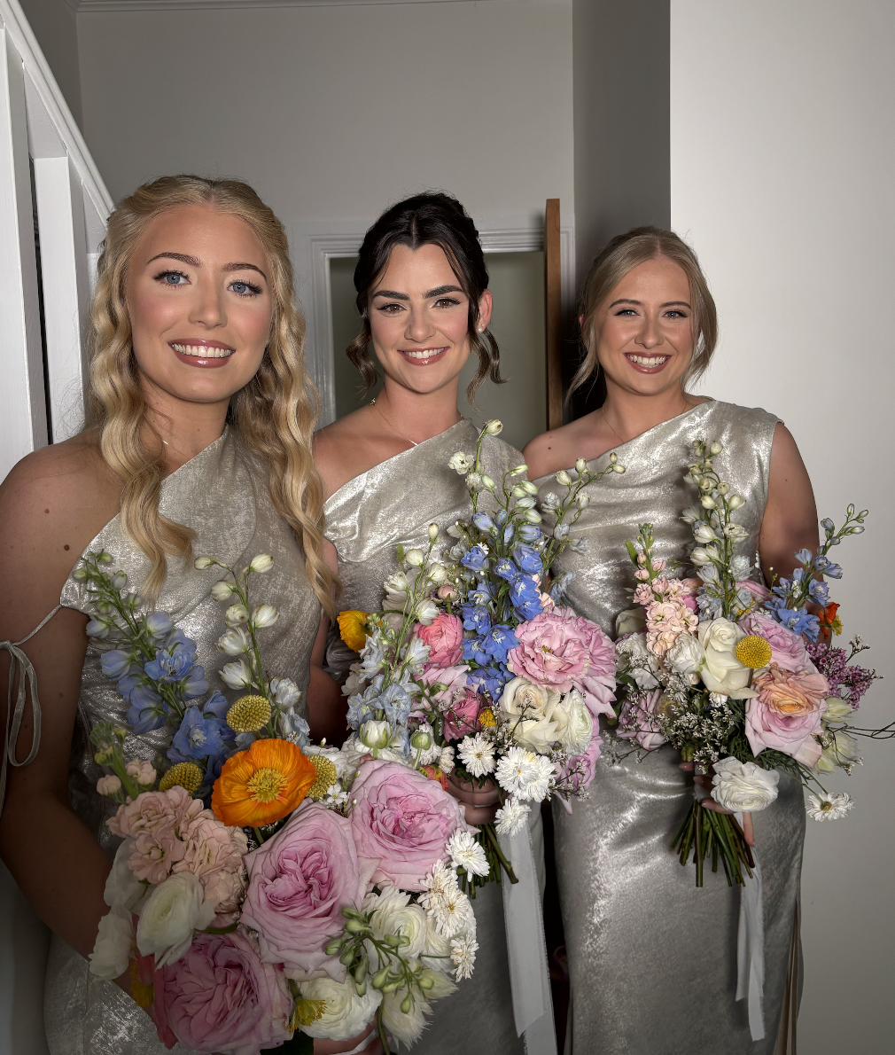 Three women in matching silver dresses holding colorful flower bouquets standing indoors in front of a wall and doorway.