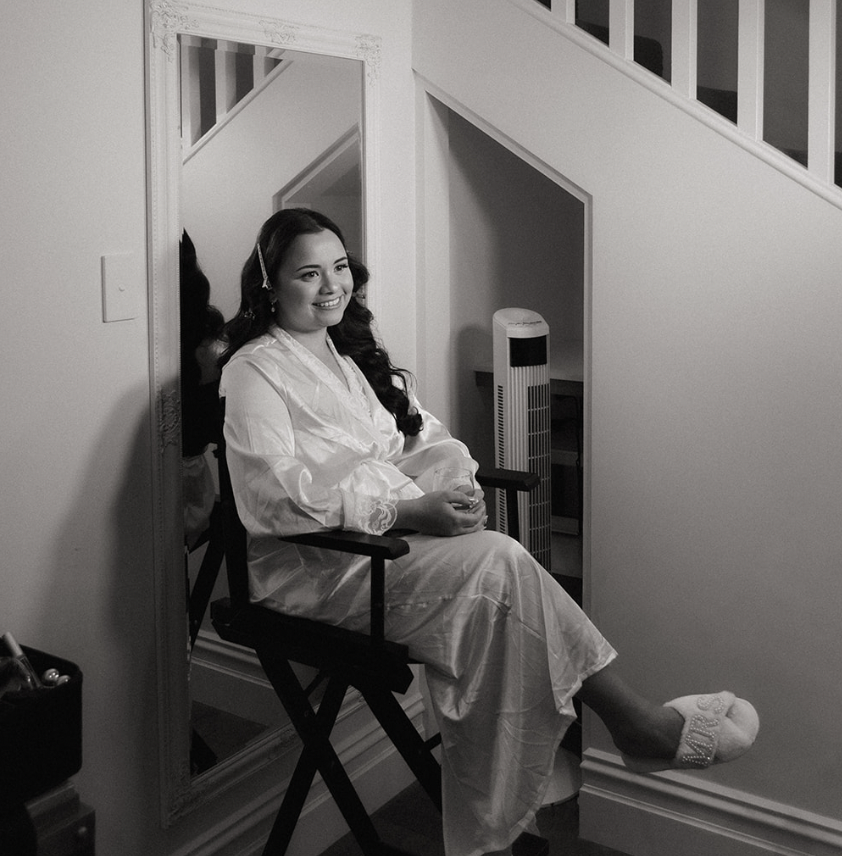 A young woman with long wavy hair sits in a director's chair, smiling, wearing a satin robe and slippers, inside a house near a staircase and a tall fan.