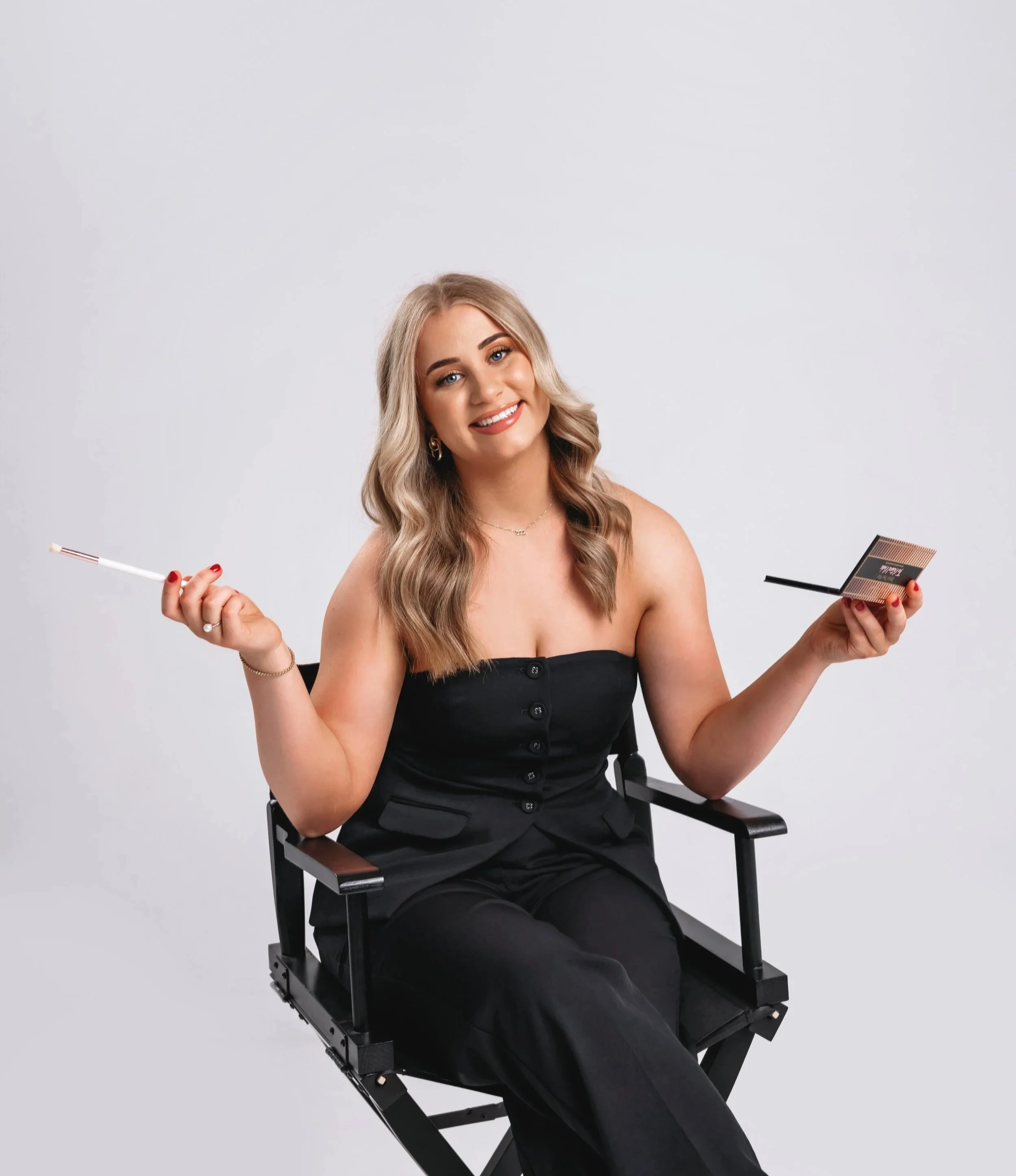 A woman with long blonde hair is sitting on a black director's chair, smiling, holding makeup tools, a brush in the right hand and a compact mirror in the left hand, against a plain light gray background.