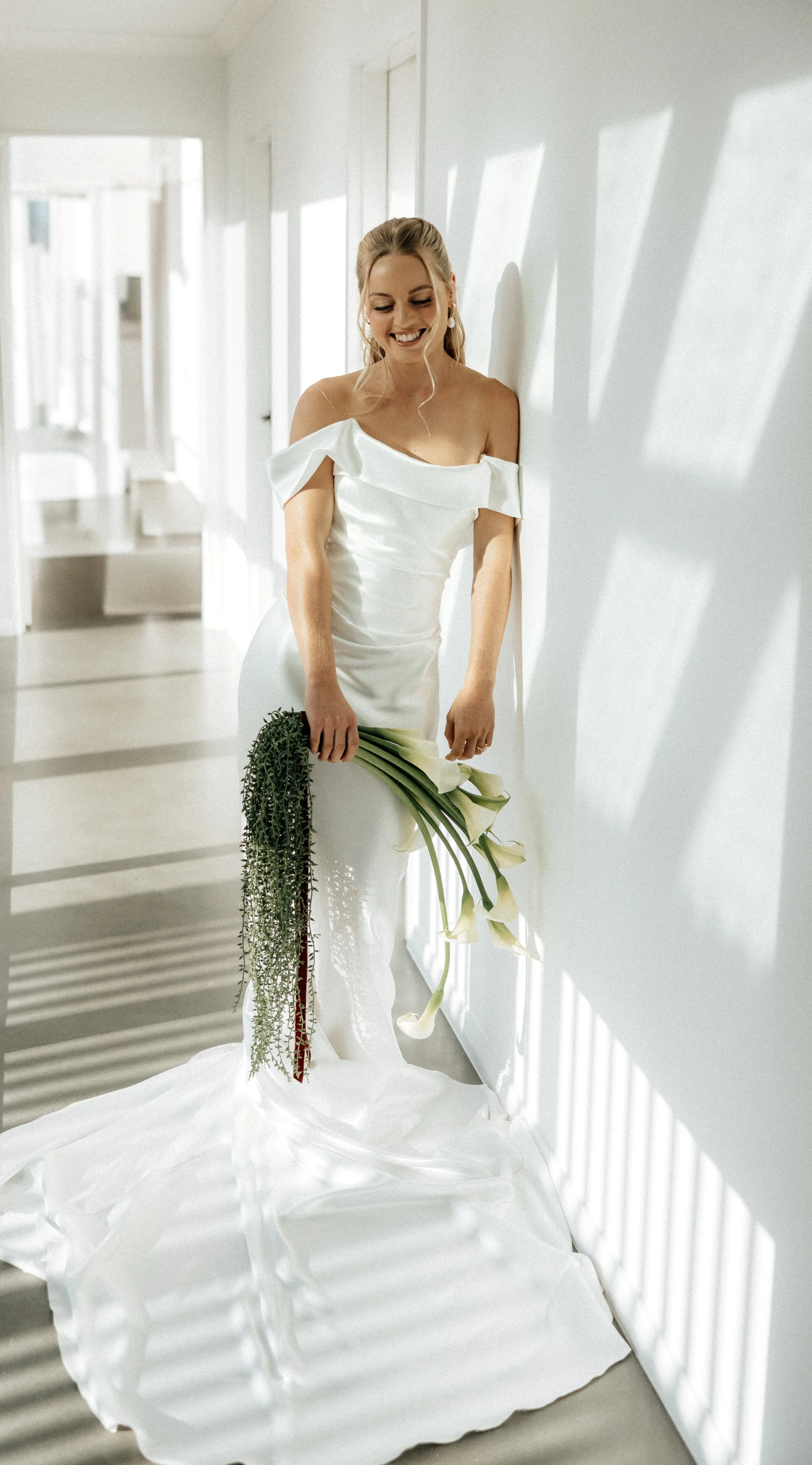 A bride in a white wedding dress standing in a sunlit room, holding a bouquet of white calla lilies, smiling and leaning against a white wall.