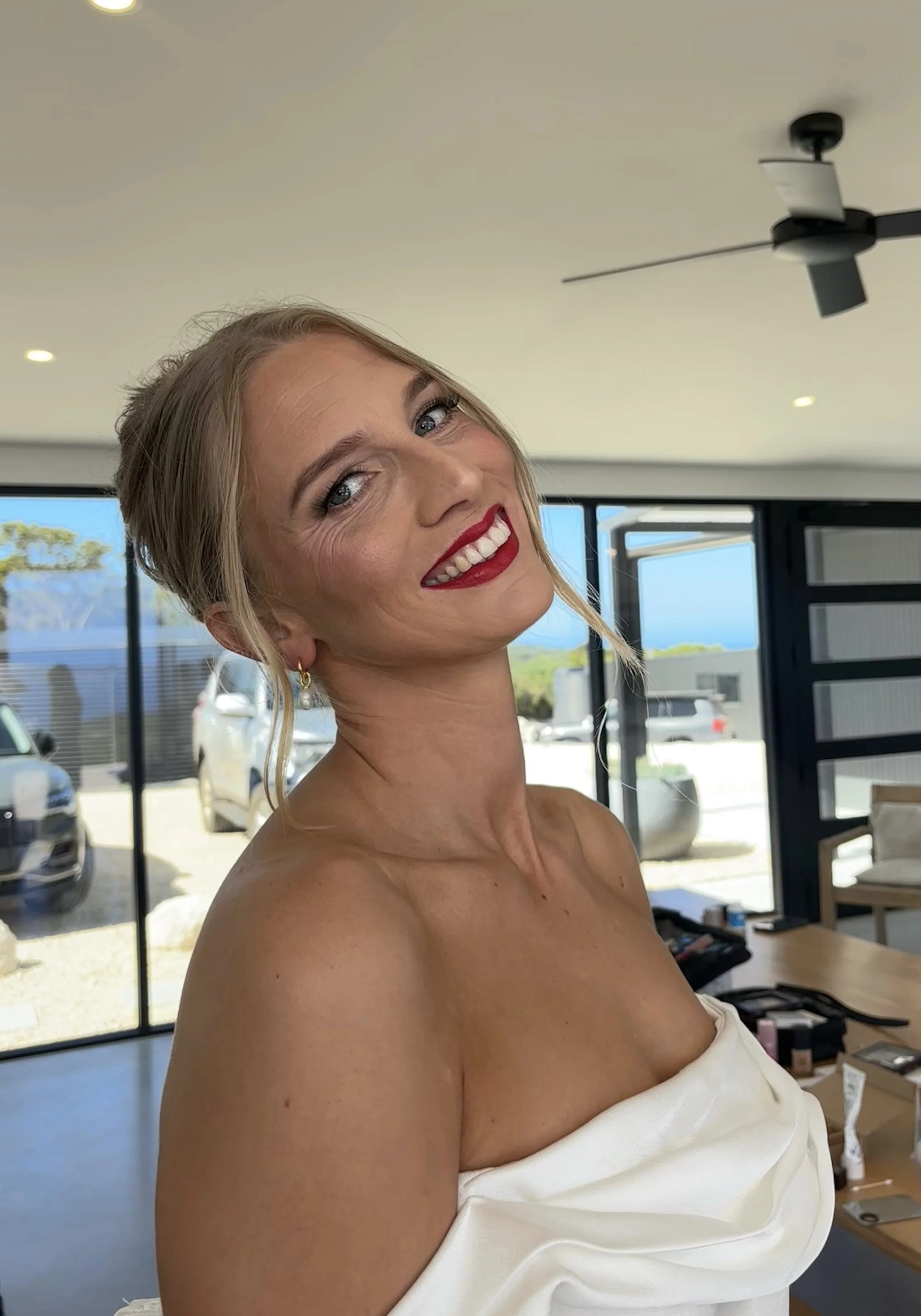 A woman with blonde hair, red lipstick, and earrings smiling at the camera indoors. She is wearing an off-shoulder white top, with cars and a bright outdoor scene visible through large windows behind her.