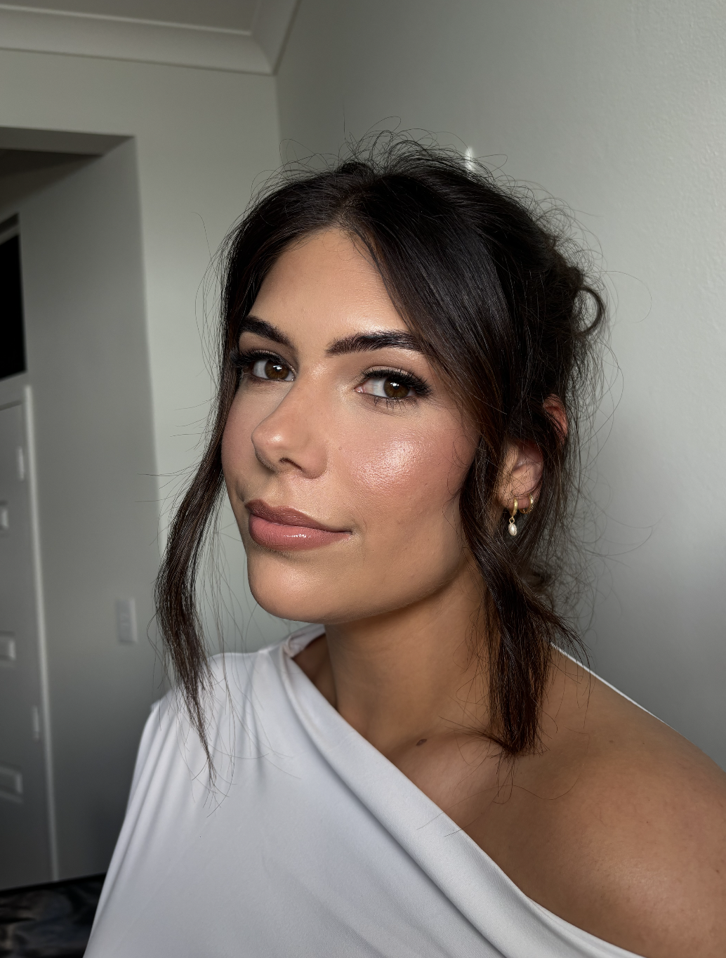 A woman with dark brown hair styled in loose curls, wearing a white off-the-shoulder top, gold earrings, and makeup, looking at the camera with a slight smile against a white wall background.