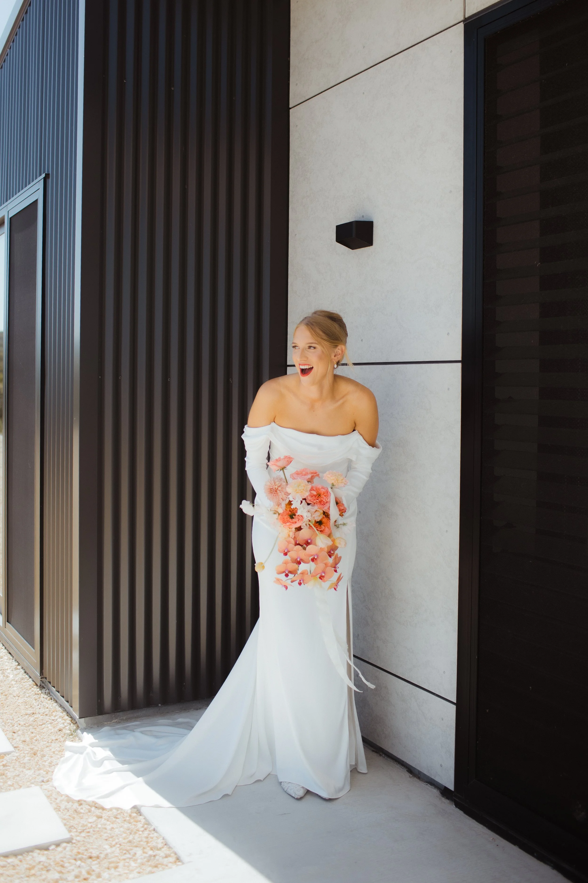 A bride in an off-shoulder white wedding dress holding a bouquet of pink and orange flowers, laughing and standing against a modern building exterior.