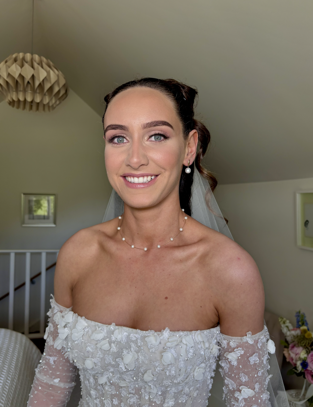 A smiling bride with blue eyes and dark brown hair styled in curls, wearing a white off-the-shoulder wedding dress with floral embroidery, a pearl necklace, matching pearl earrings, and a wedding veil, standing in a room with soft lighting.