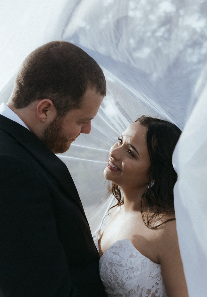 A bride and groom on their wedding day, standing under a sheer white fabric, looking into each other's eyes and smiling.