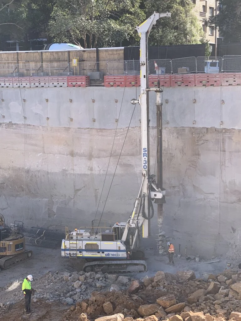 Construction site with a large drill machine drilling into the ground, two workers wearing safety vests and helmets supervising, rocks and dirt gathered around, and buildings and trees in the background.