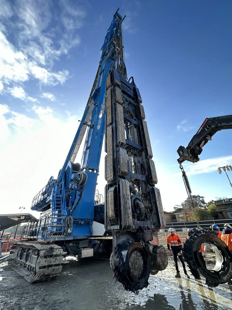 A large blue construction crane with a long boom arm lifting large industrial parts. Several construction workers wearing safety helmets and orange vests are on the ground nearby.