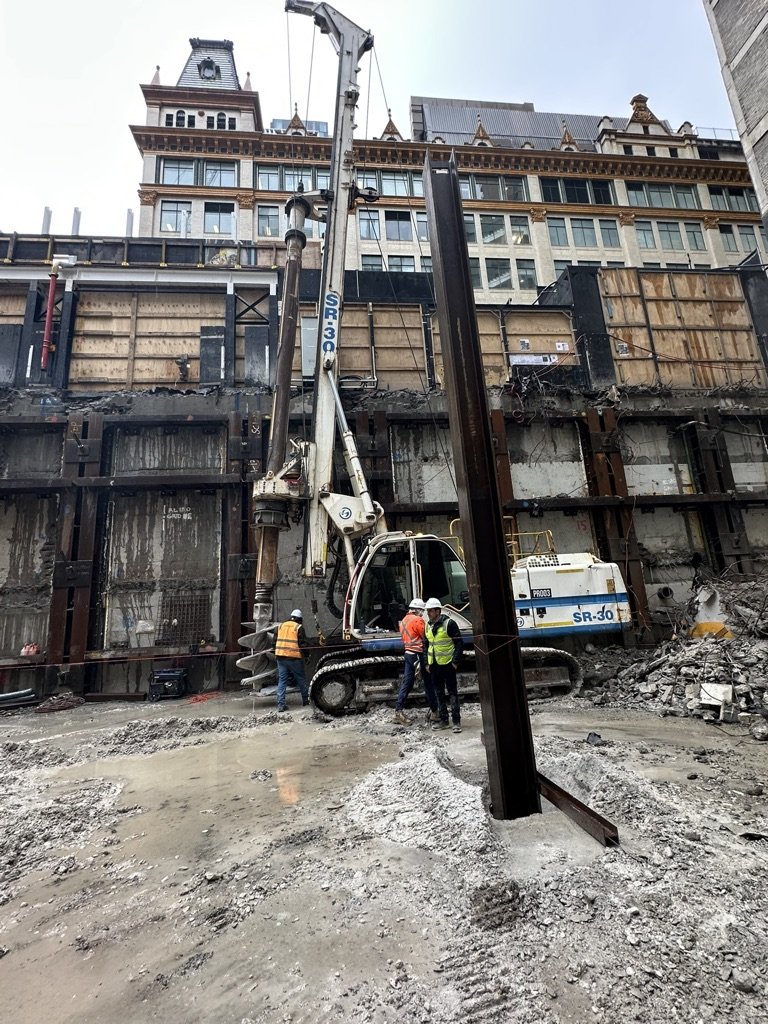 Construction workers operating a drilling or piling machine at an urban construction site with existing multi-story building in the background.