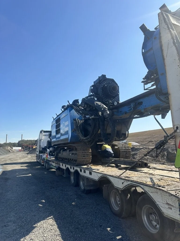 A large construction machine, possibly an excavator, being transported on a flatbed truck at a construction site under a clear blue sky.