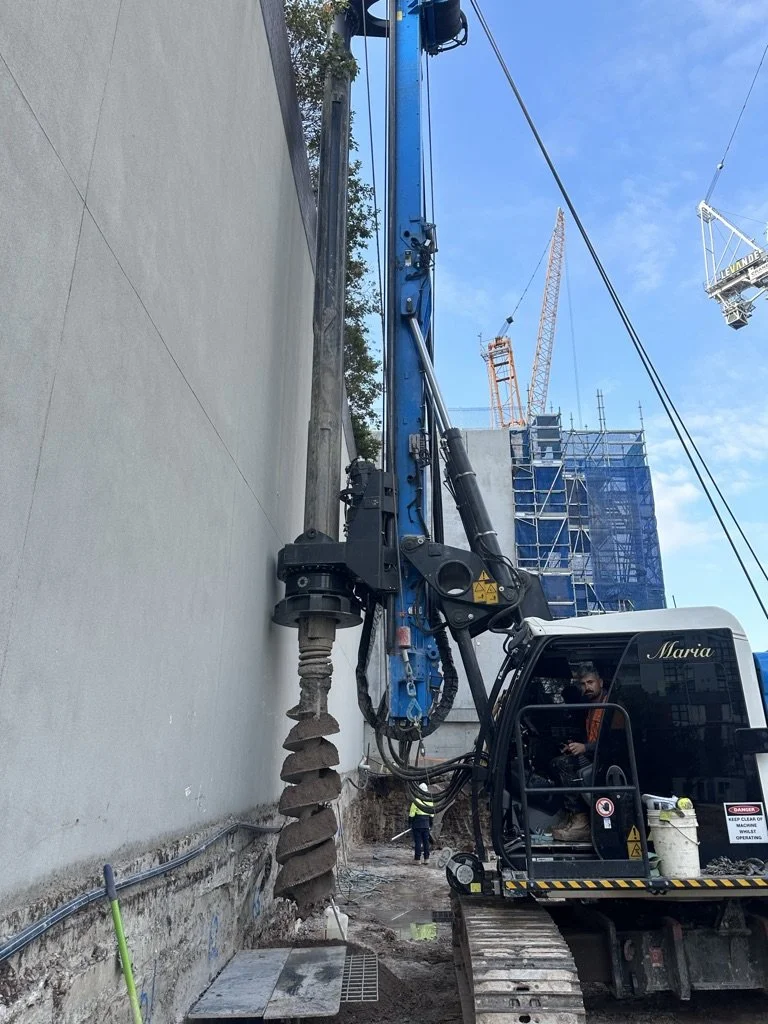 Construction site with a large blue drill attached to a hydraulic excavator, drilling into the ground beside a concrete wall, with construction workers and high-rise buildings in the background.
