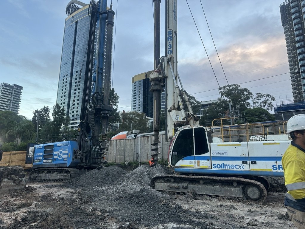 Construction site with excavation equipment and workers, with tall modern buildings in the background.