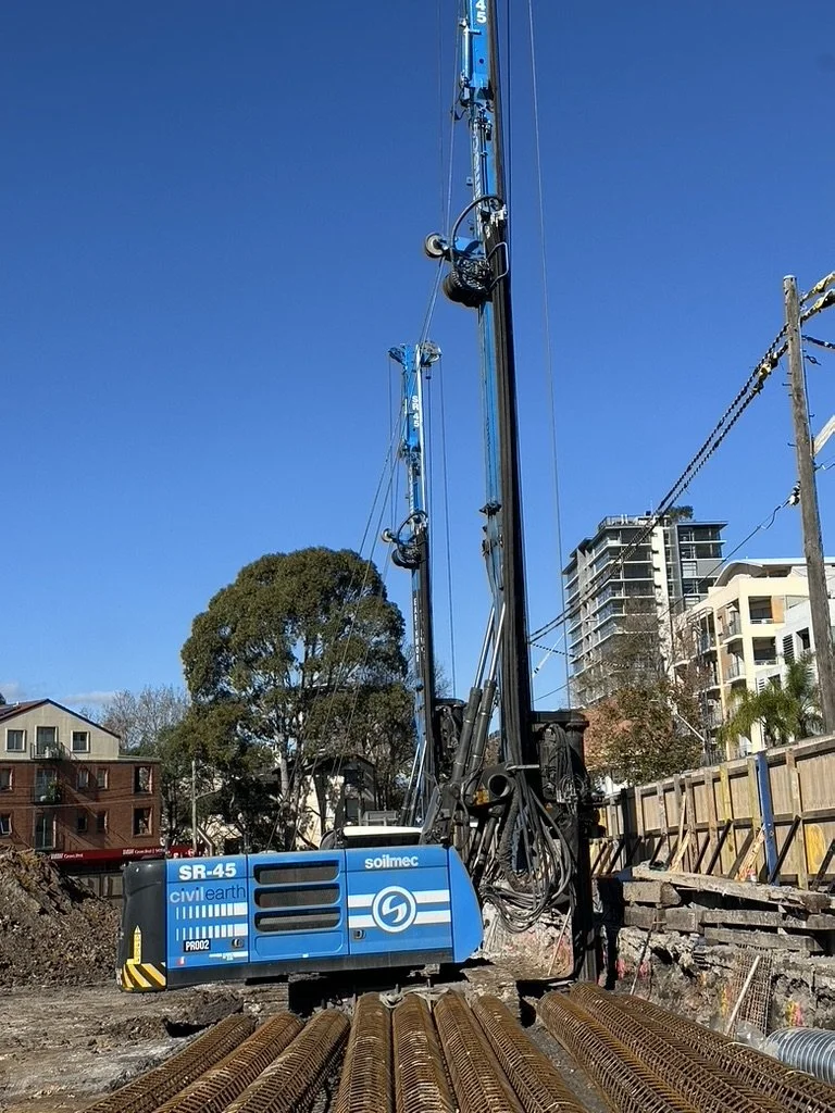 A large blue and black soil drilling machine at a construction site with steel reinforcement bars in the foreground and urban buildings in the background under a clear blue sky.