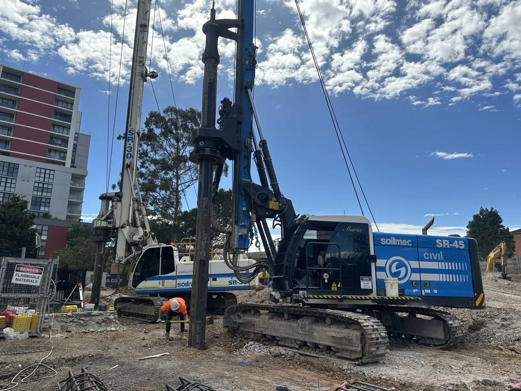 Construction site with large drilling equipment and a worker in an orange safety vest and helmet, with residential buildings and a partly cloudy sky in the background.