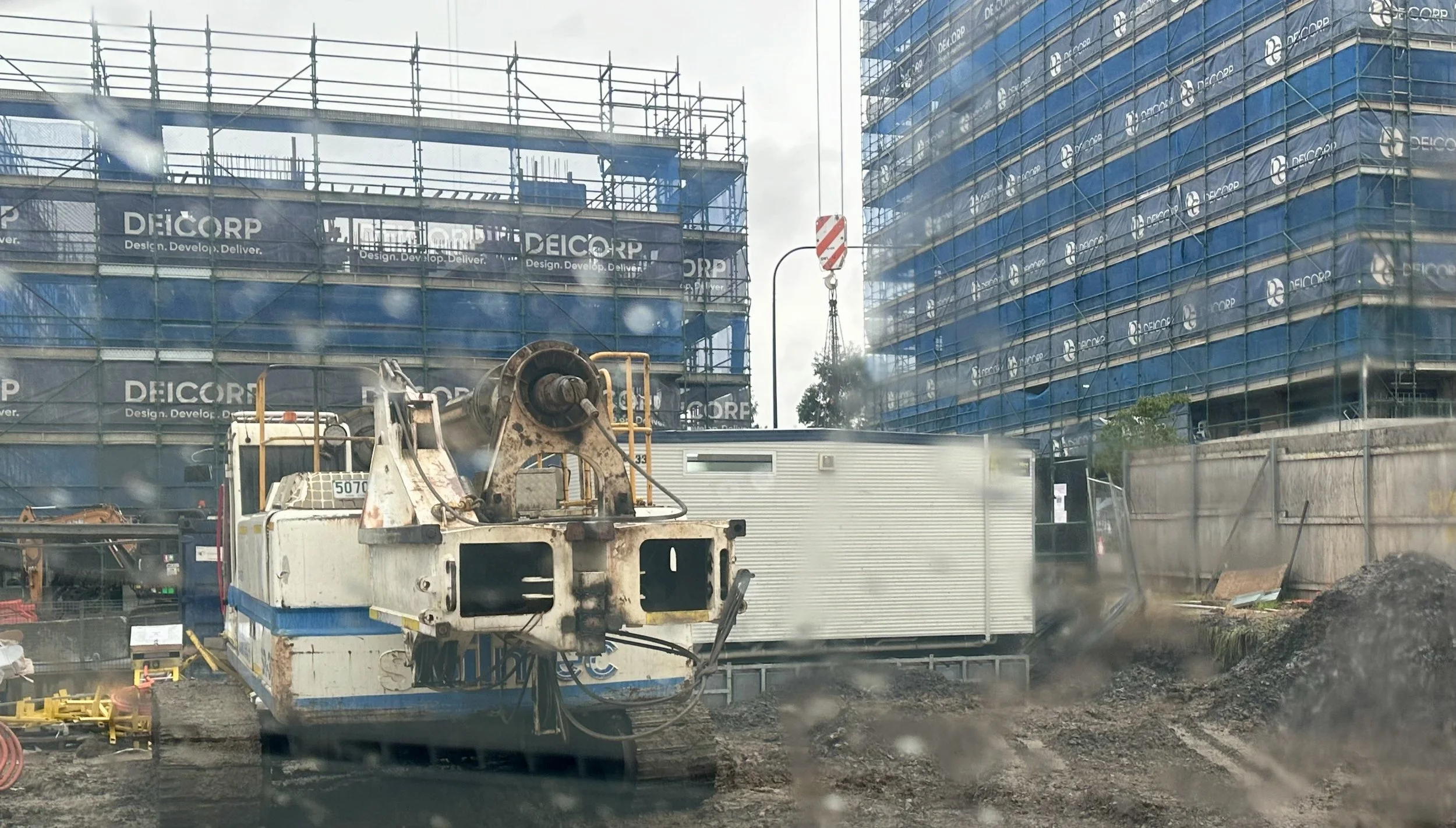 Construction site with two buildings covered in blue scaffolding and a crane in the background. In the foreground, there's construction equipment and muddy ground, viewed through a dirty window.