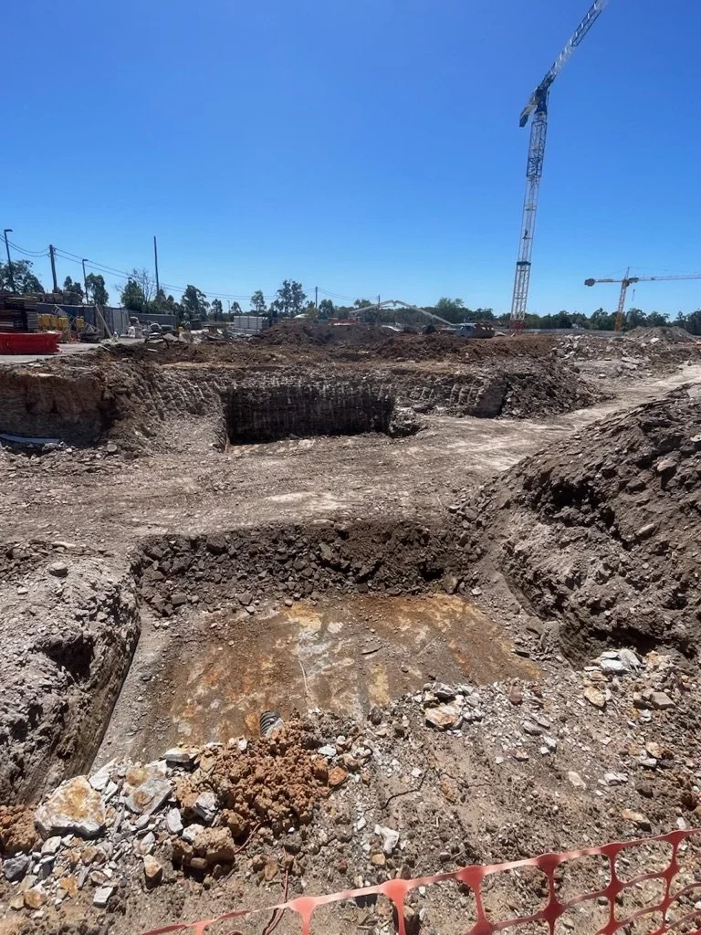 Construction site with large excavated area, dirt, rocks, and construction cranes in the background under a clear blue sky.