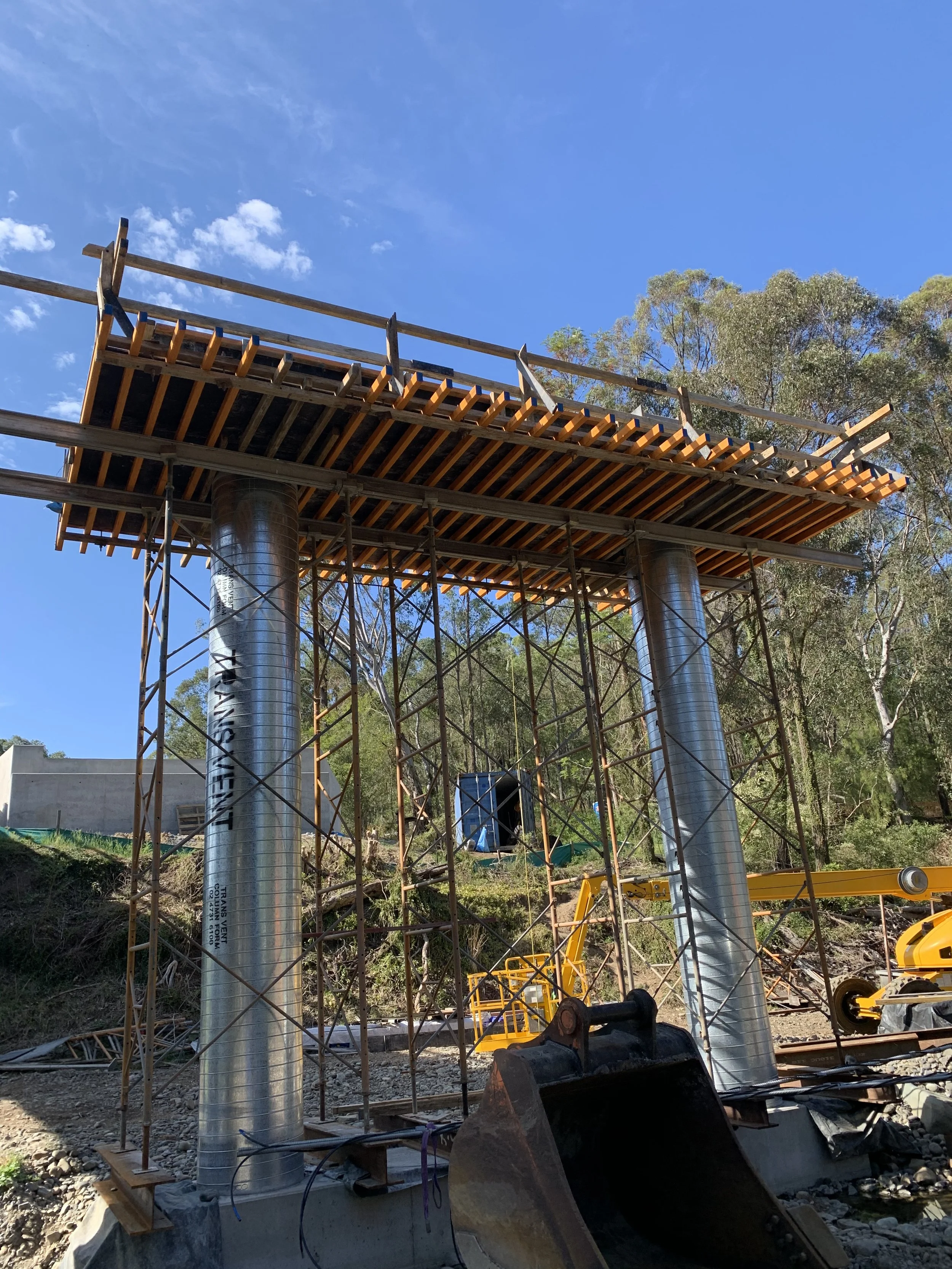 Under construction steel bridge with scaffolding and construction equipment, trees in the background, blue sky.