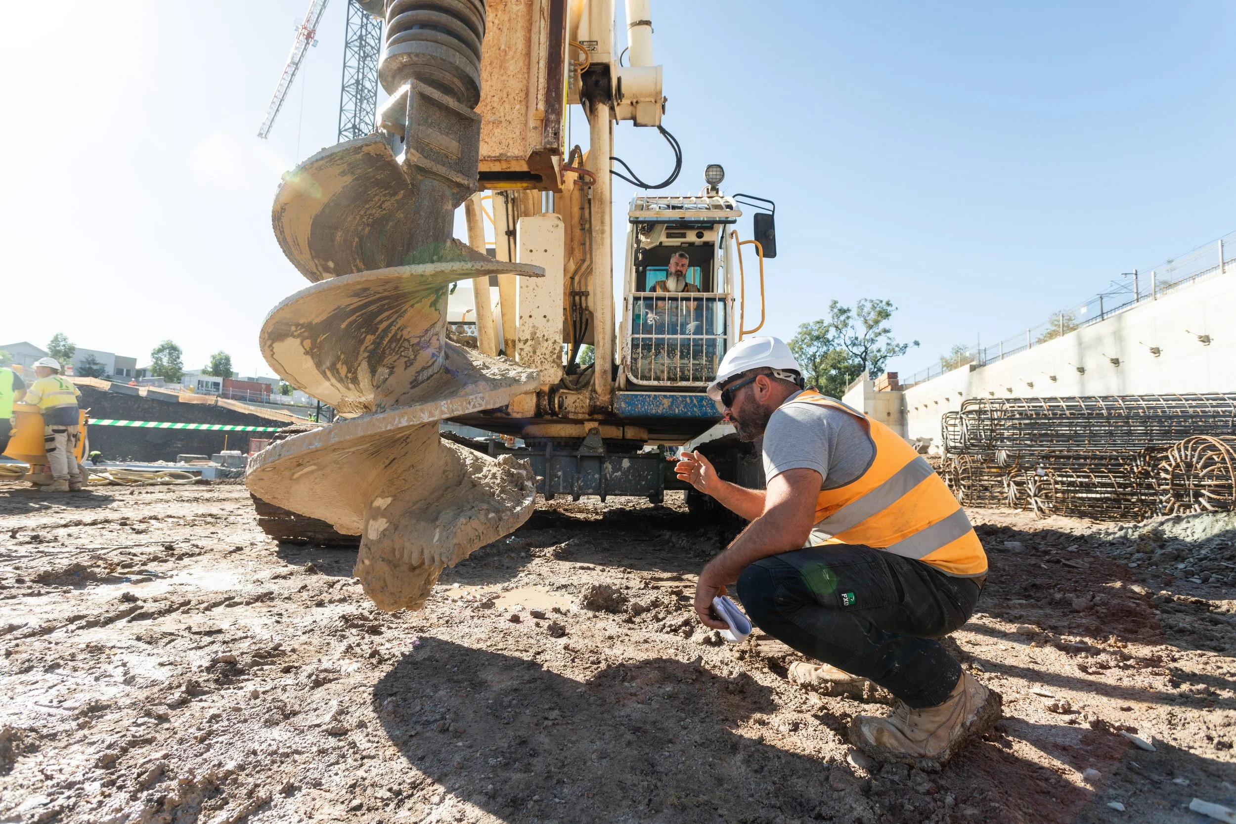Construction worker crouching near a large yellow drilling machine on a construction site, with a man operating the machinery in the background, and other workers visible in the distance.