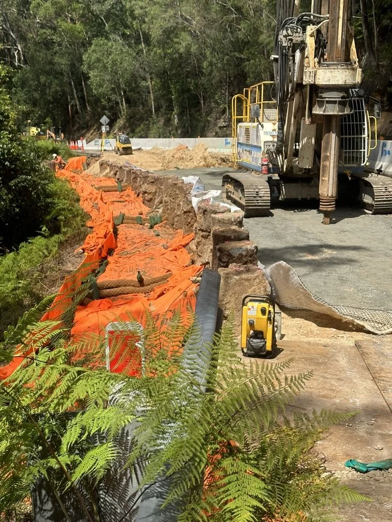 Construction site with an excavator working on a road, orange safety barriers, sandbags, and construction equipment, with greenery and trees in the background.