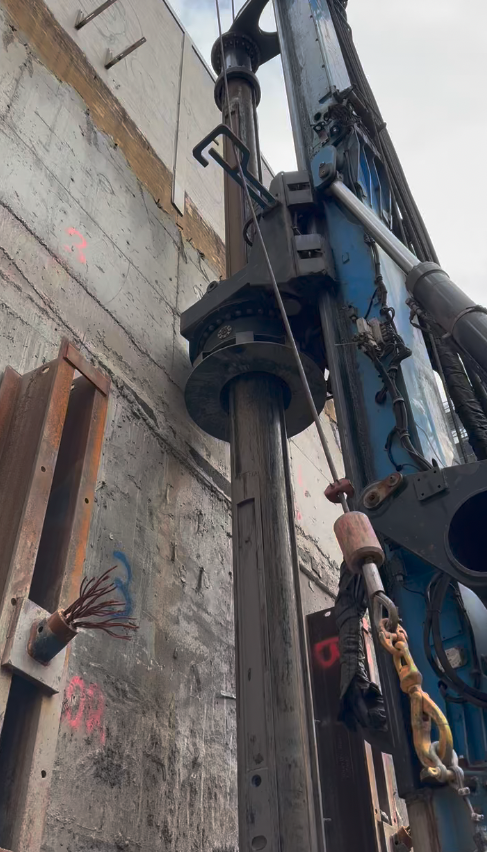 Close-up view of a large hydraulic piling or drilling machine positioned next to a concrete wall at a construction site.